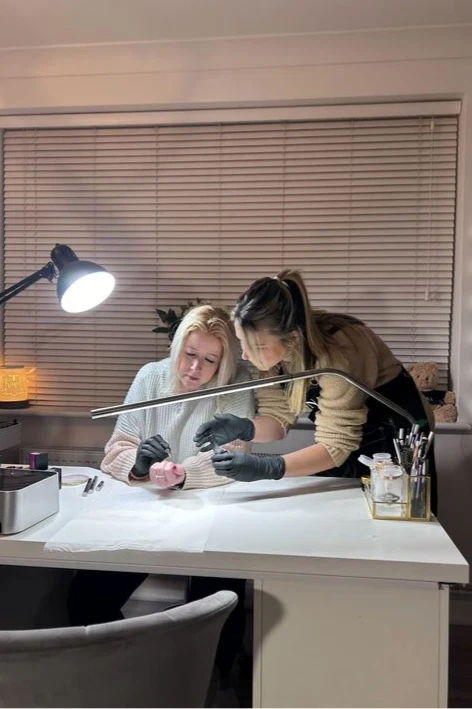 Two women working together at a desk, one doing nail art.