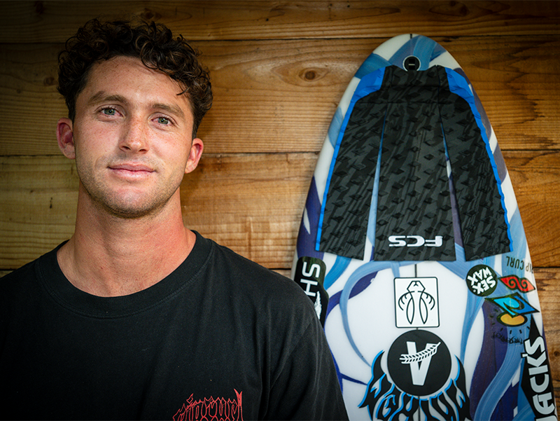 Person smiling beside a surfboard against a wooden background.