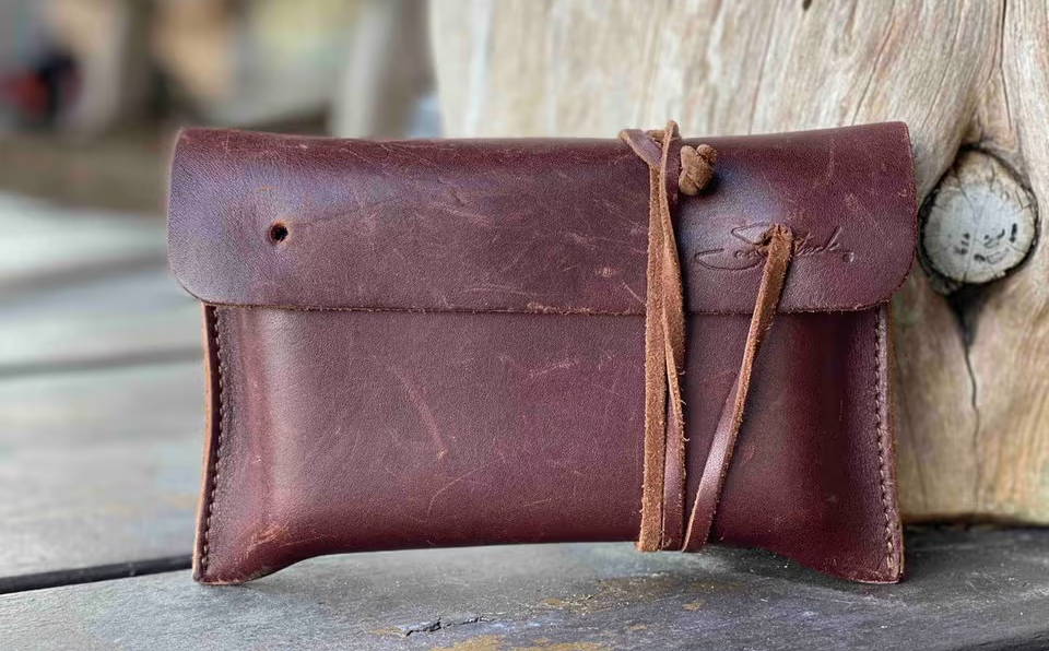 Dark brown full-grain leather wallet with natural scuffs and a tied leather cord leaning against rustic weathered wood.