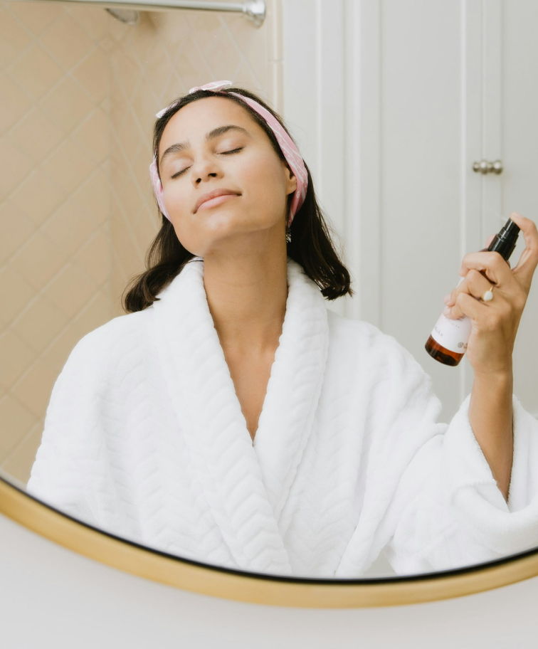 A woman in a bath robe stood in a bathroom, spraying herself with moisturizer
