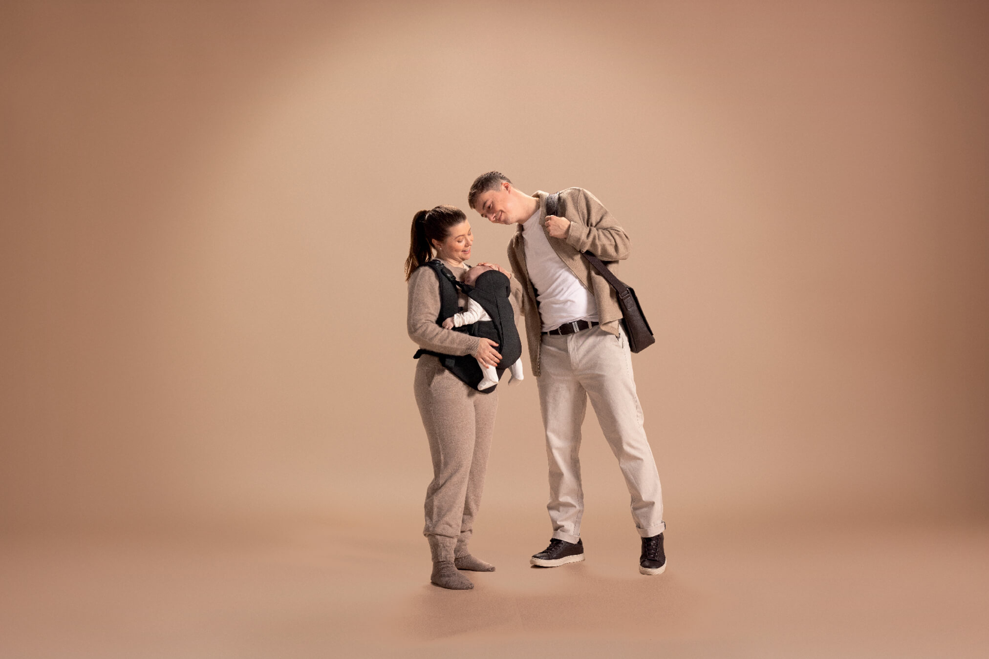 A couple holding a baby with a Kudo baby carrier in a neutral-colored setting.