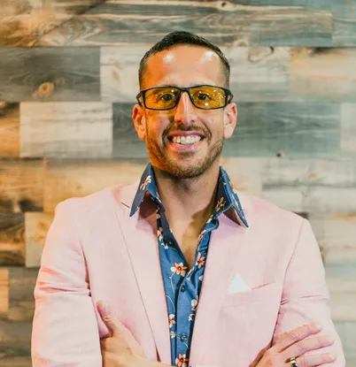 Smiling man in a pink blazer and yellow glasses against a wooden background.