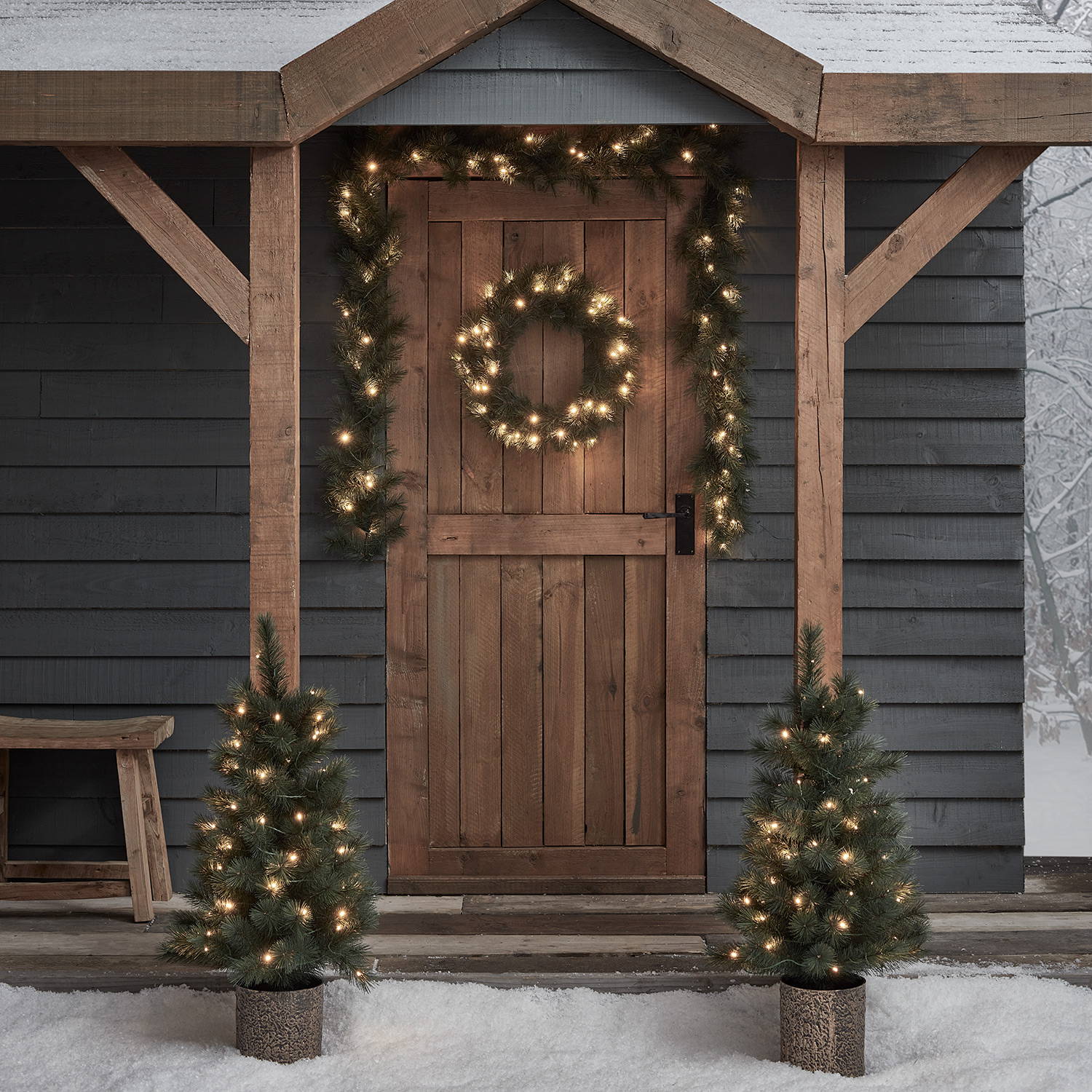 A wintery porch decorated with faux green wreath, garland and mini trees, each illuminated by warm white LEDs
