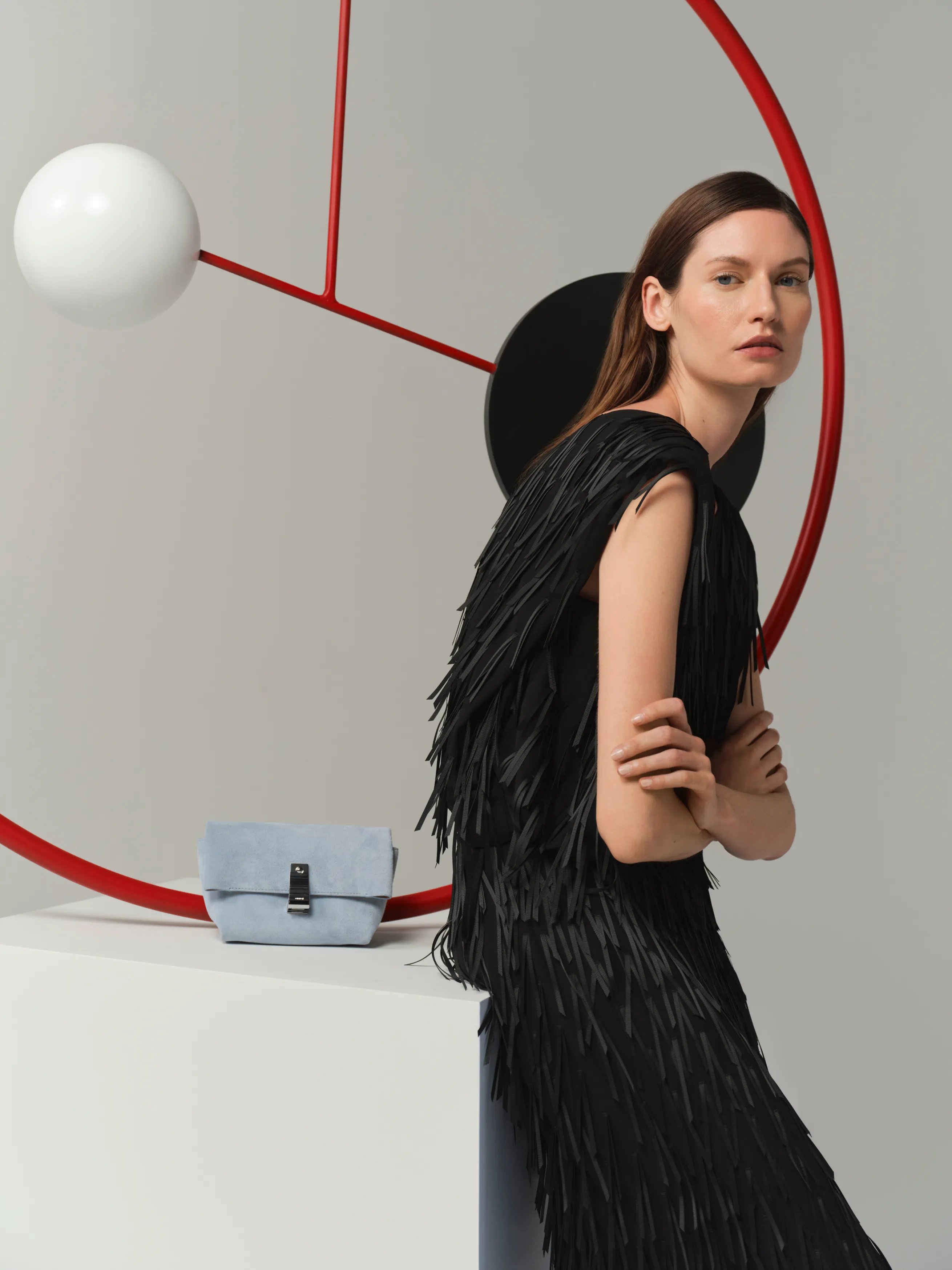 Model in a black fringed dress poses with a blue handbag and geometric background.