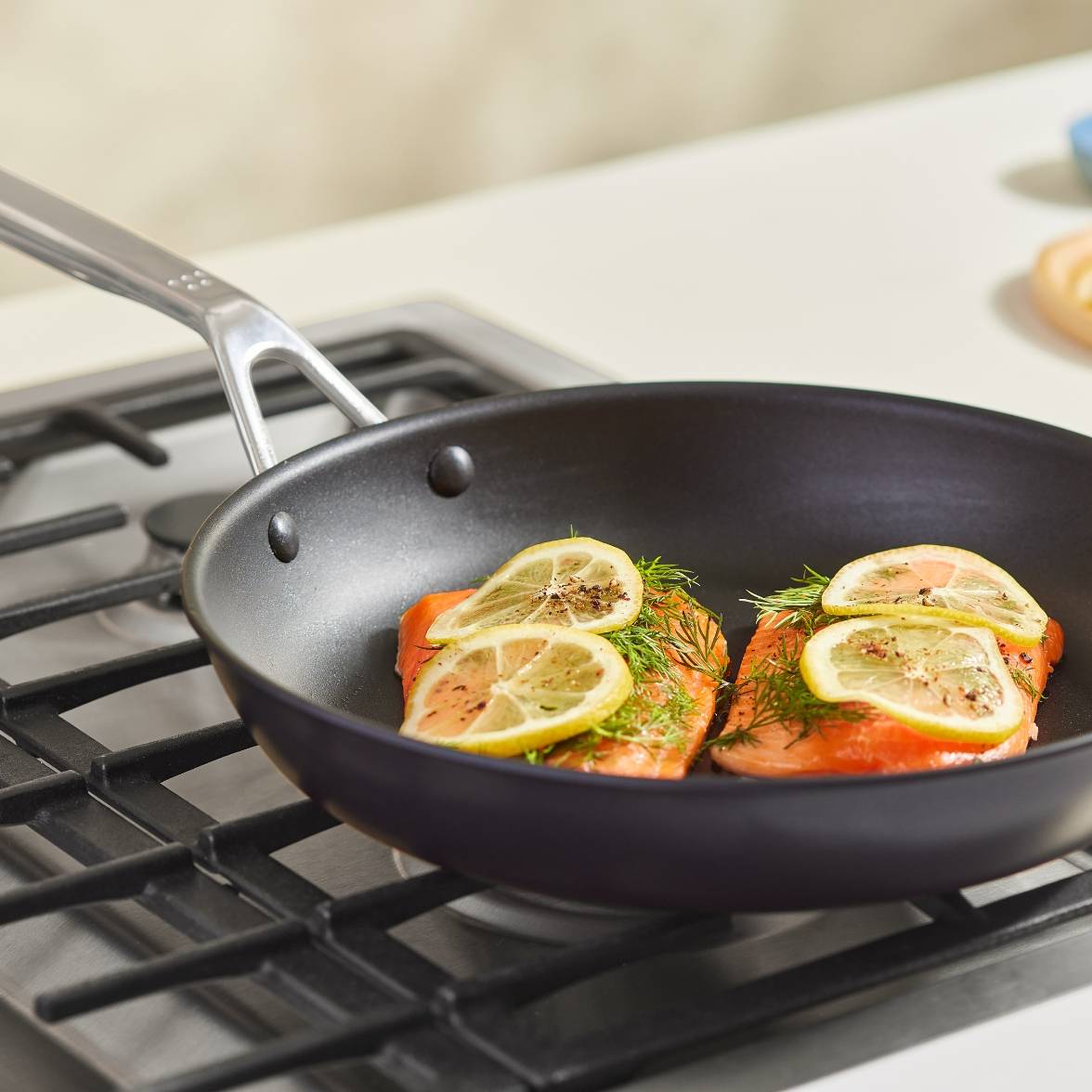 Two salmon filets topped with herbs and sliced lemon sit in a Misen Nonstick Pan on a stovetop.