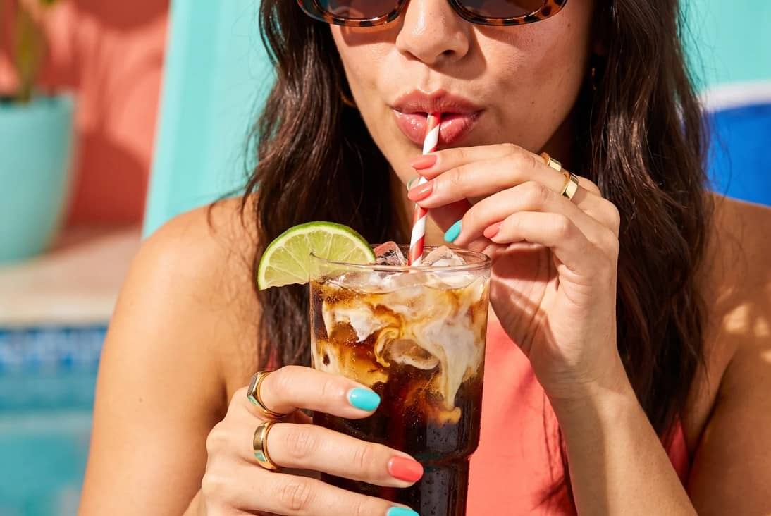 A woman in sunglasses sips a cola drink with a lime garnish through a striped straw poolside