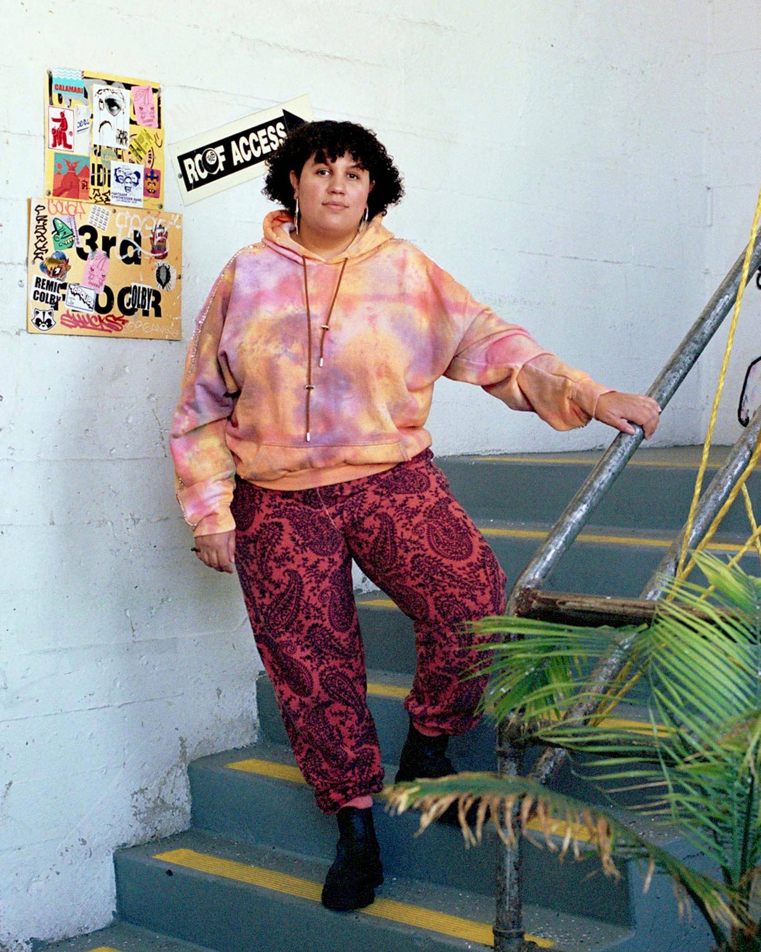 Image of Sara Ekua Todd, standing in a stairwell, wearing a tie dye sweatshirt and paisley sweatpants. 