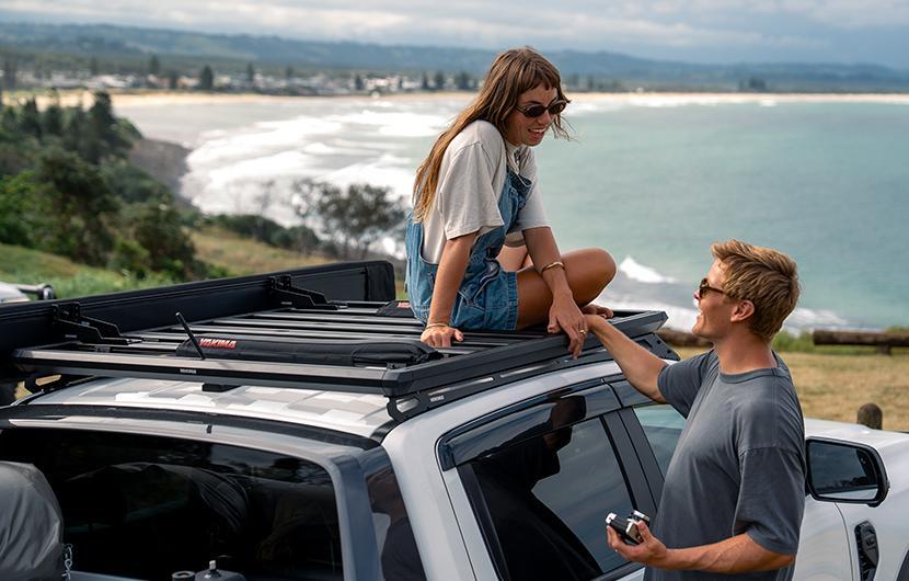 A woman sitting on top of a roof rack platform, with her partner next to her in front of a beach