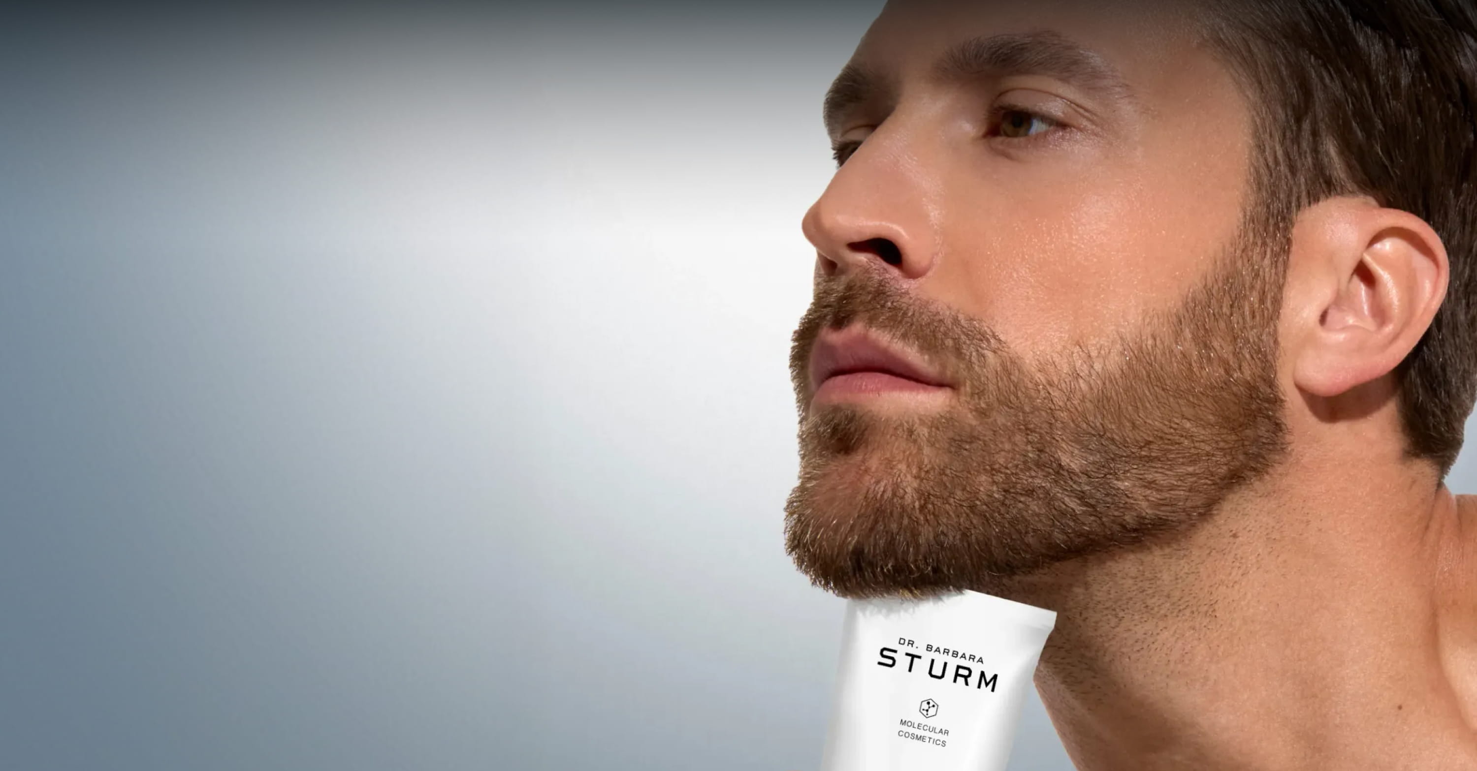 Man with a beard looking upward, product tube visible in the background.