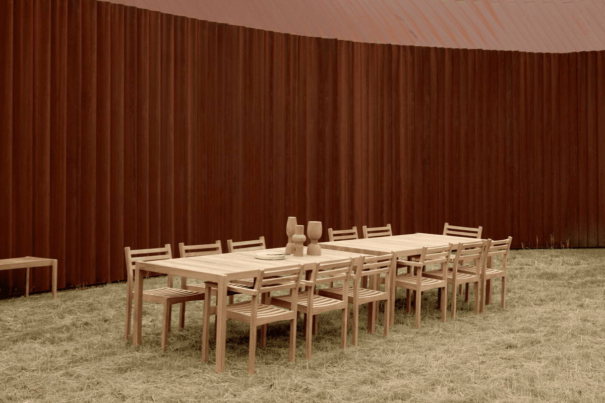 Boxhill's Alfred Outdoor Teak Dining Table and Chairs set up banquet style in the grass against a curved brown wall.