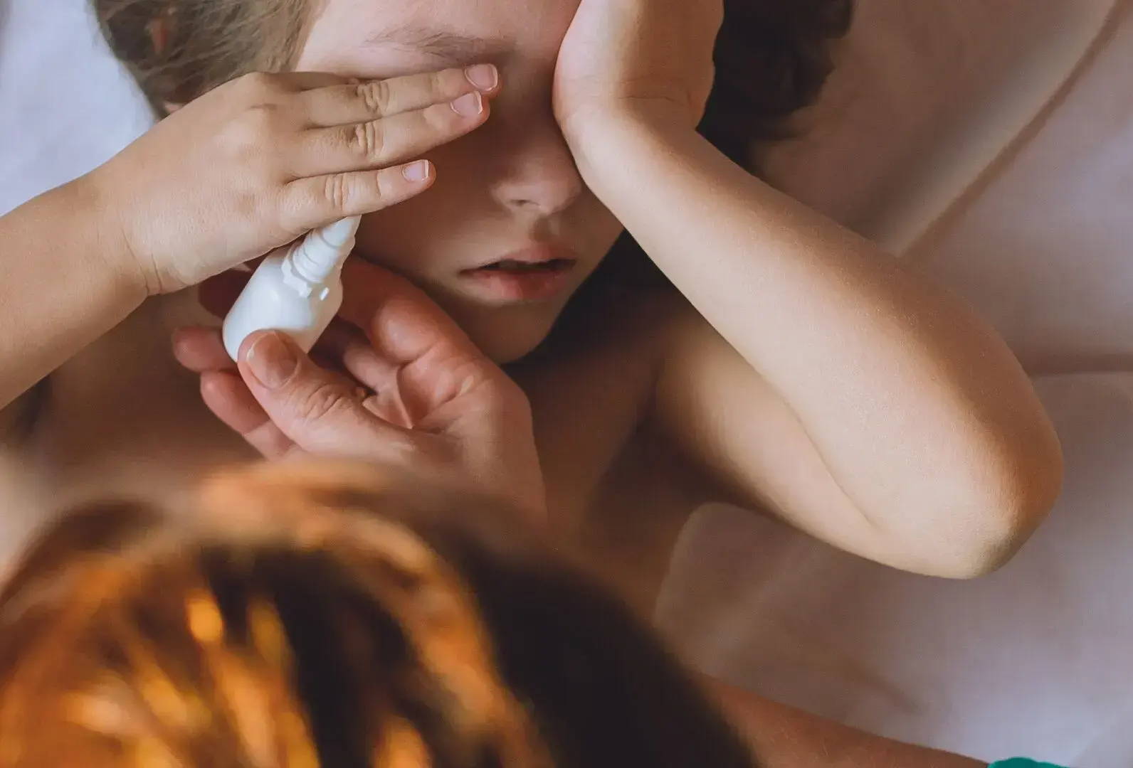 Dievča leží v posteli s rukami na tvári, mama sa nad ňu nakláňa a dáva jej očné kvapky na upokojenie svrbenia z dôvodu alergie