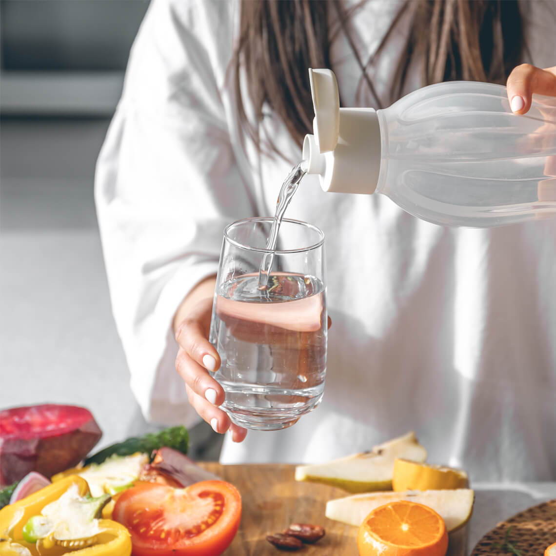 Person pouring water into a glass with fruits on a wooden table.
