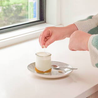 A hand adding an ingredient to a dessert on a plate by a window.