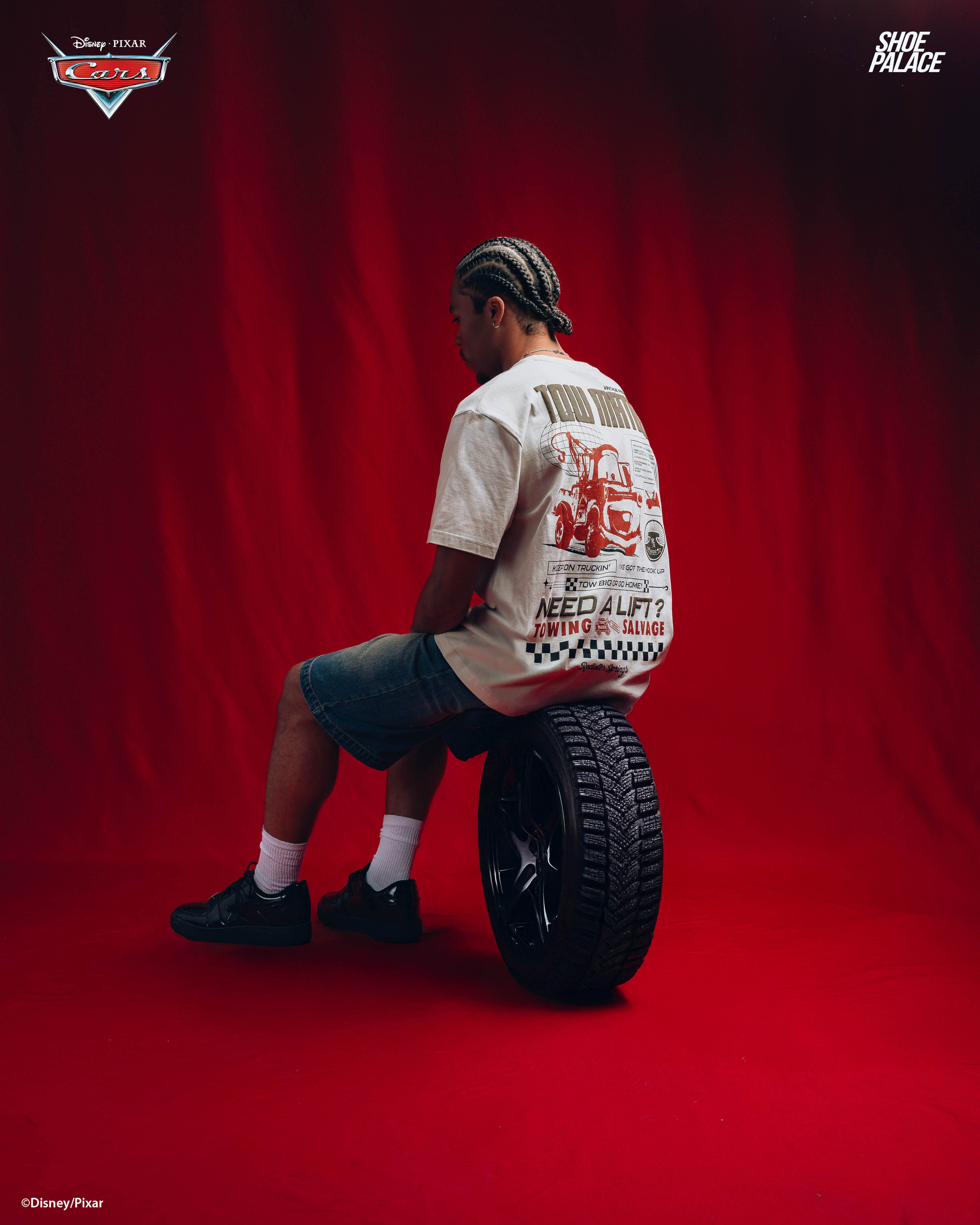 Person sitting on a tire against a red backdrop, wearing a graphic t-shirt.