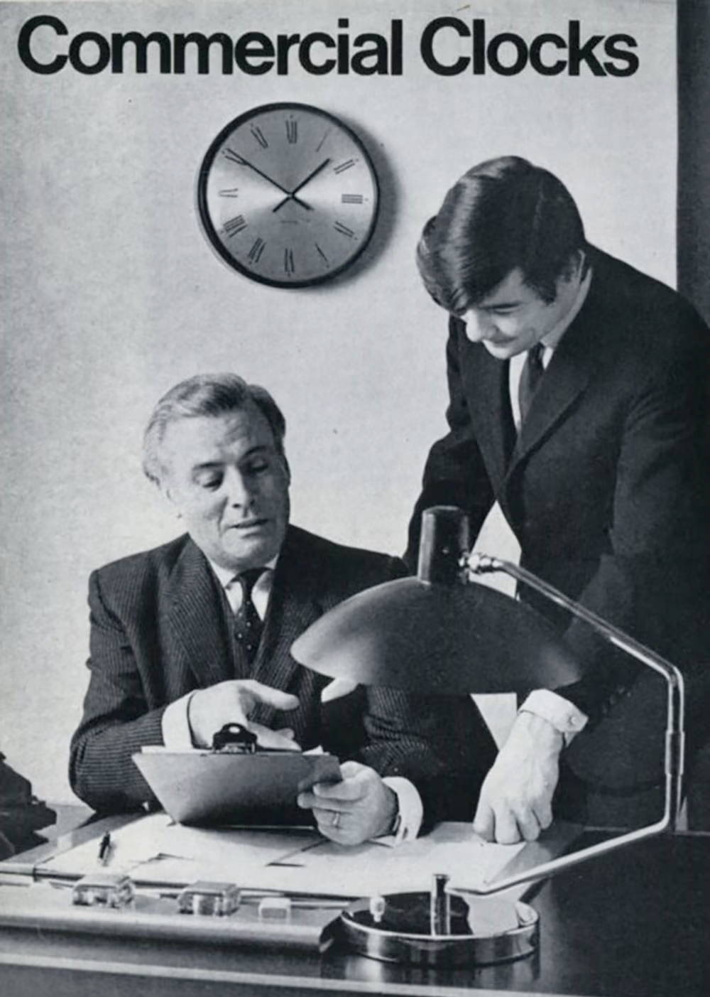 Two men discussing documents at a desk, with Westclox Manager 12-24 Hour clock in the background.