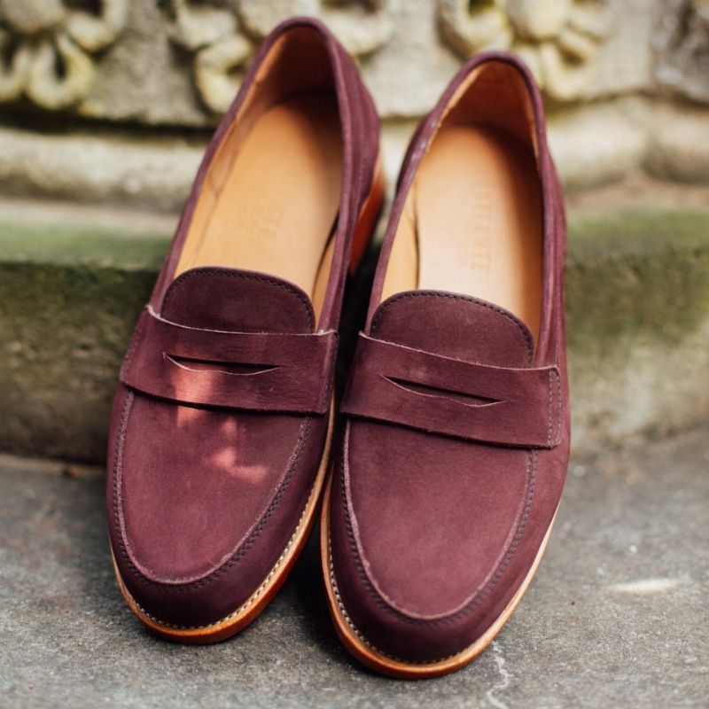 A top-down lifestyle view of the handcrafted Adelante Sofia women's wide width classic penny loafers in a rich plum burgundy suede, resting on a stone surface.
