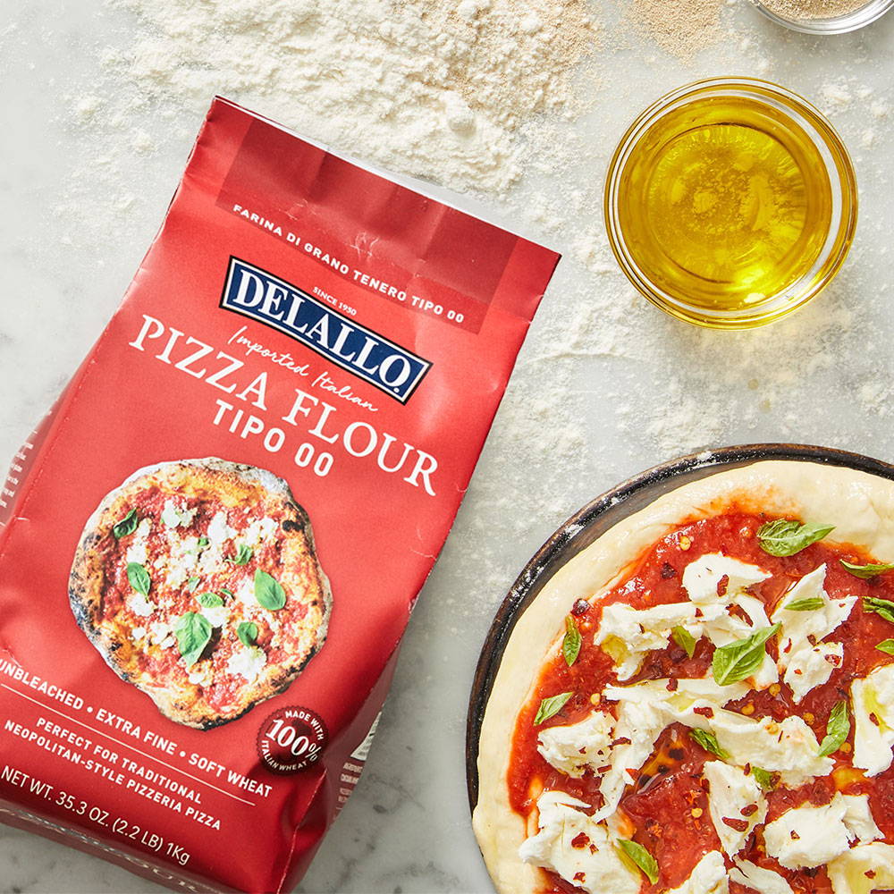 Package of DeLallo Pizza Flour laying on marble table next to raw pizza