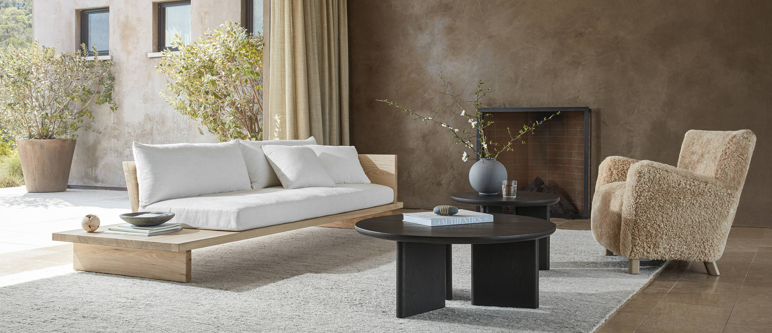 A living room furnished with a Muir sofa in Alabaster performance chenille and natural ash finish, a large and medium Morro table in Charcoal, and a Perry chair in Sandstone shearling.