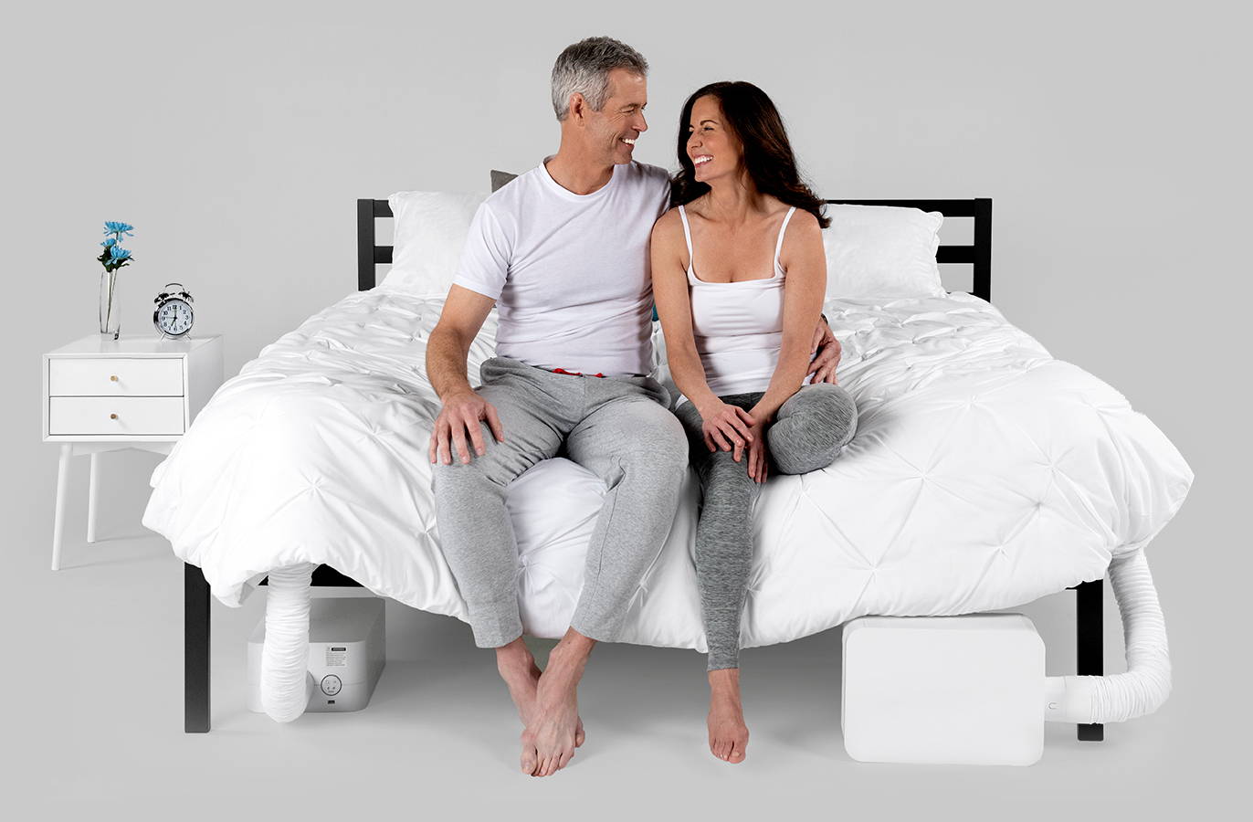An image of an older couple sitting on the edge of the bed, looking at each other and smiling, and two BedJet units are hooked up to the bed, one on each side.