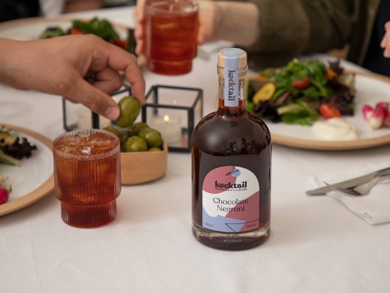 Chocolate Negroni bottle on dinner table with food in background and people picking up olives