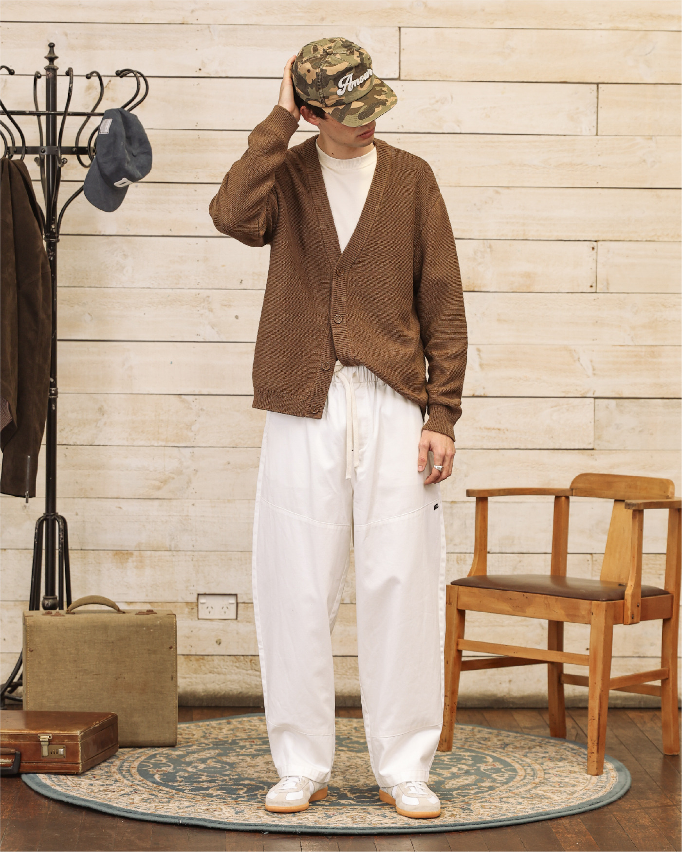 Person wearing a brown cardigan and camouflage hat, standing in a room.