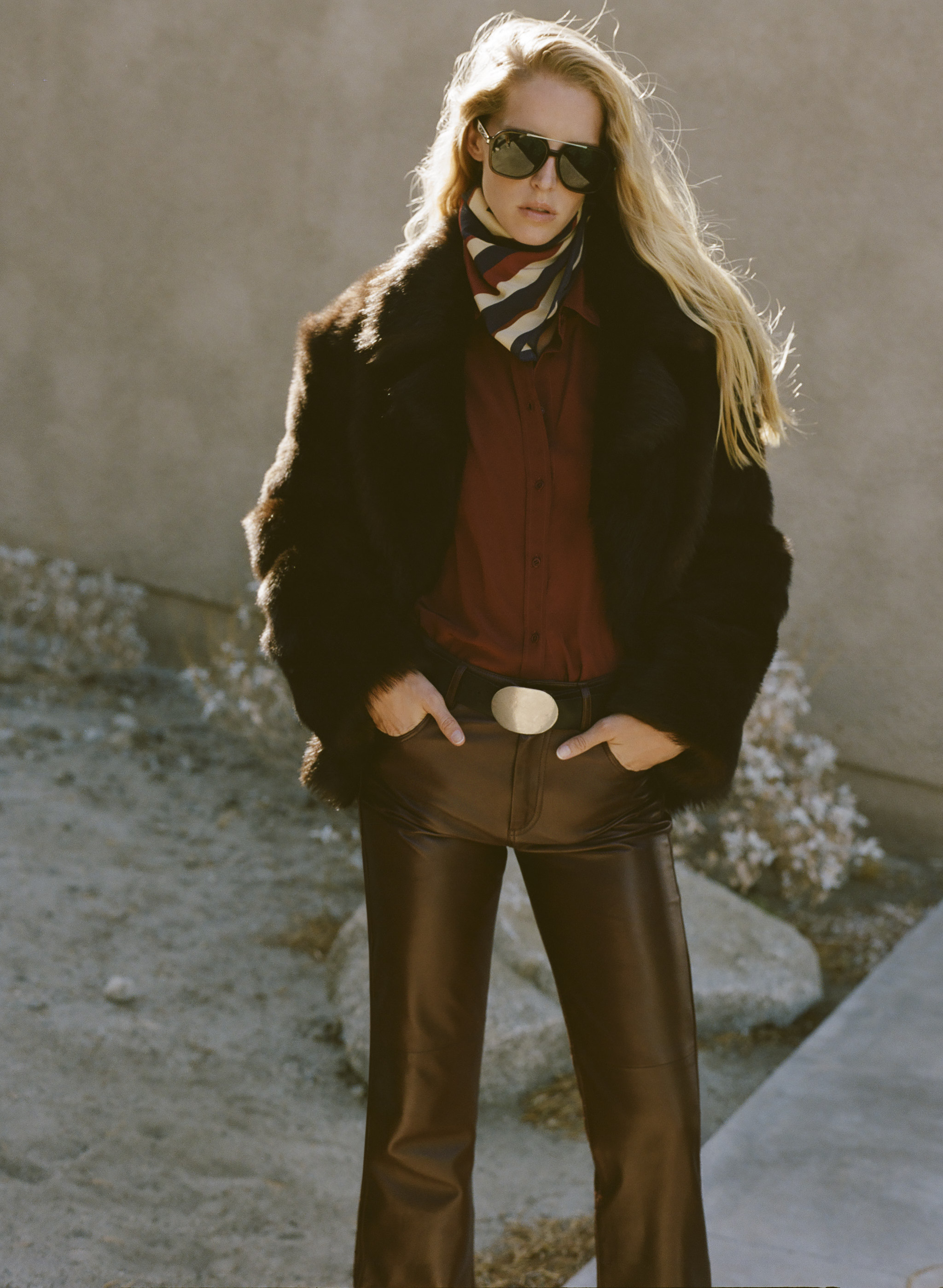 A woman in sunglasses wears a fur coat and brown pants, standing confidently.