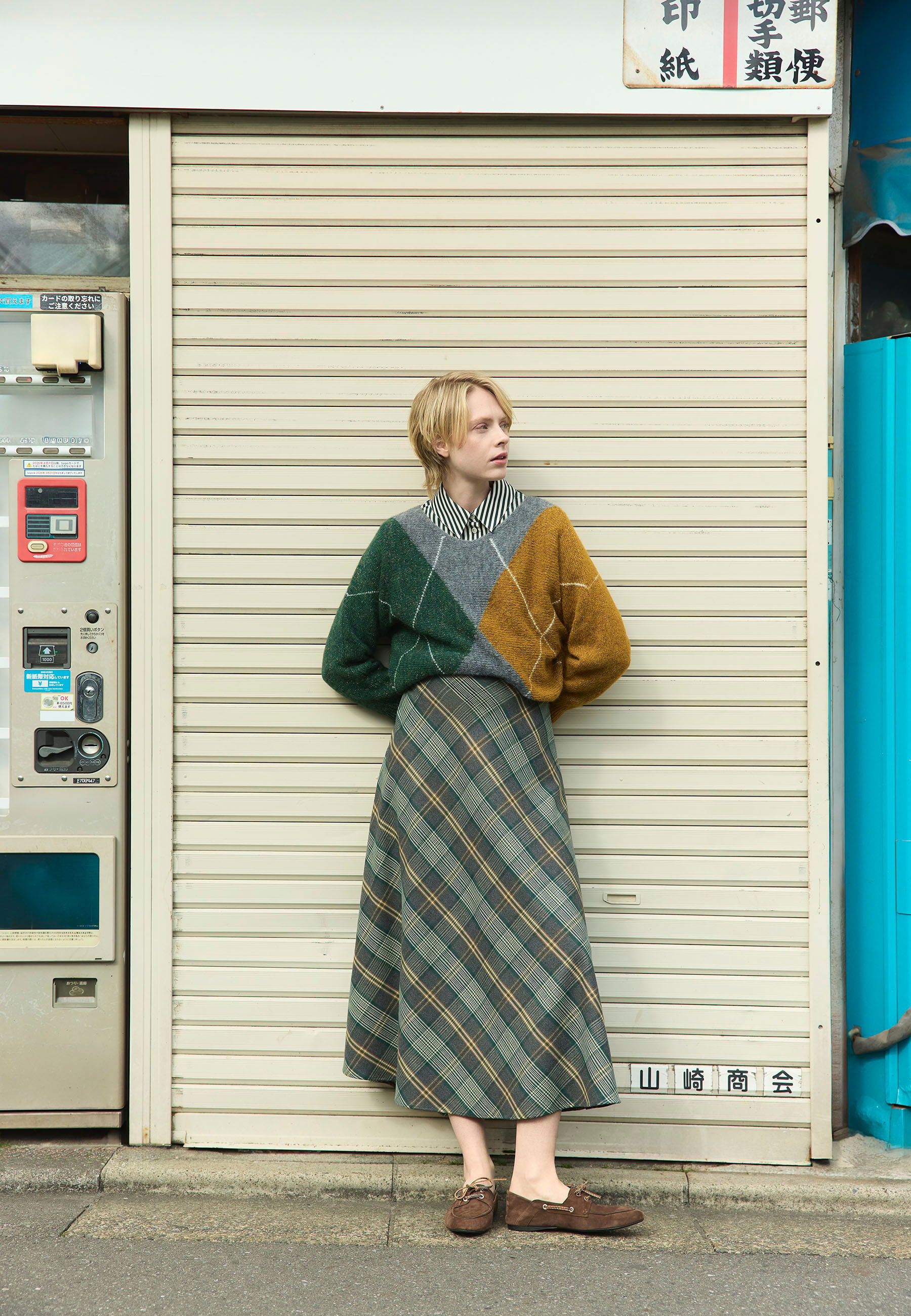A person in a colorful sweater and plaid skirt stands by a closed shop.