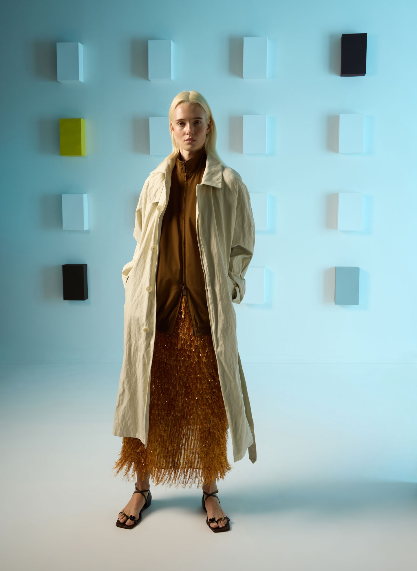Model in a long coat and fringed skirt against a minimalist background.