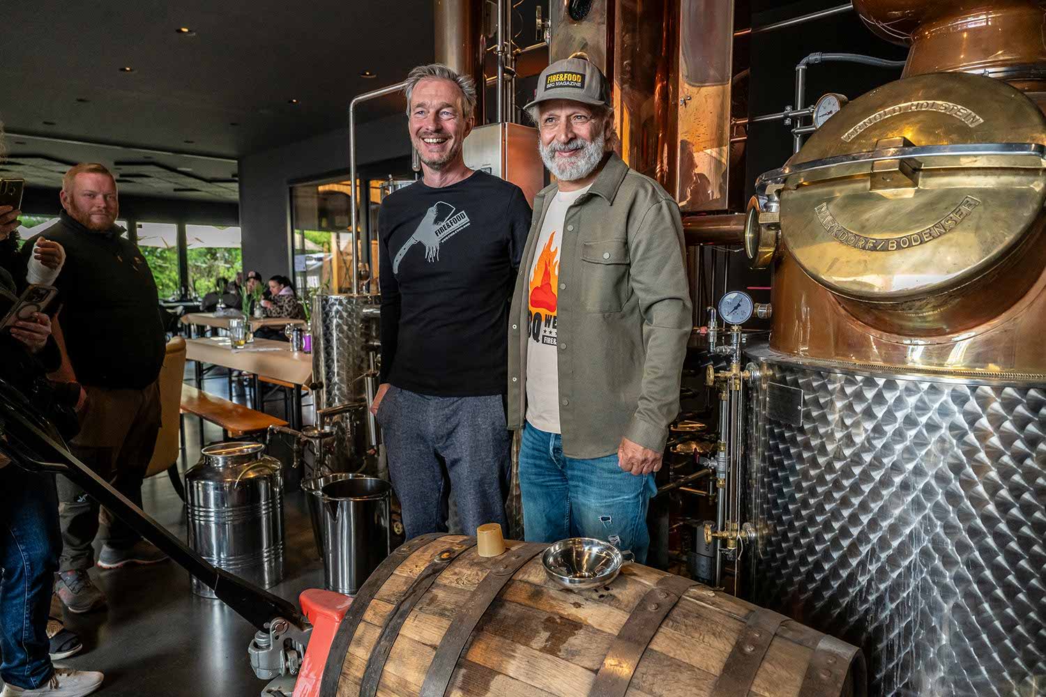 Two men posing near a distillery equipment with a barrel in the foreground.