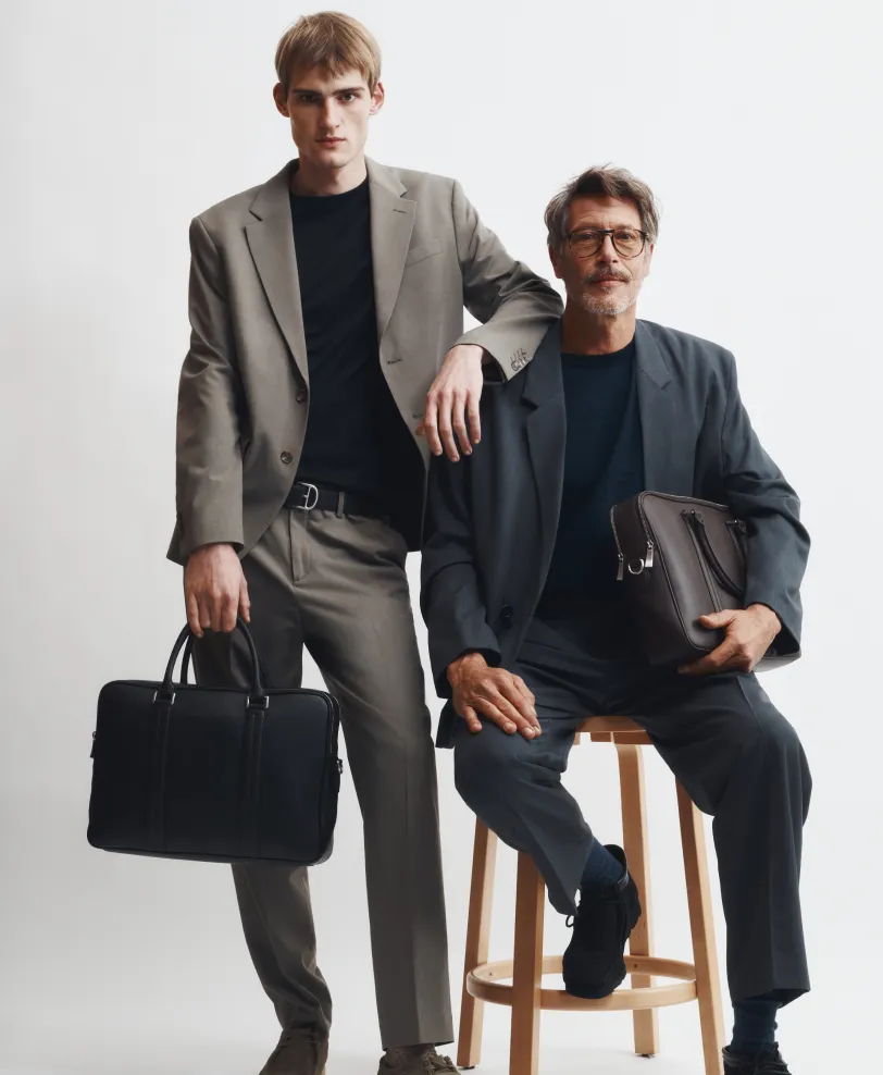 Two men in suits, one standing and one seated, both holding bags.
