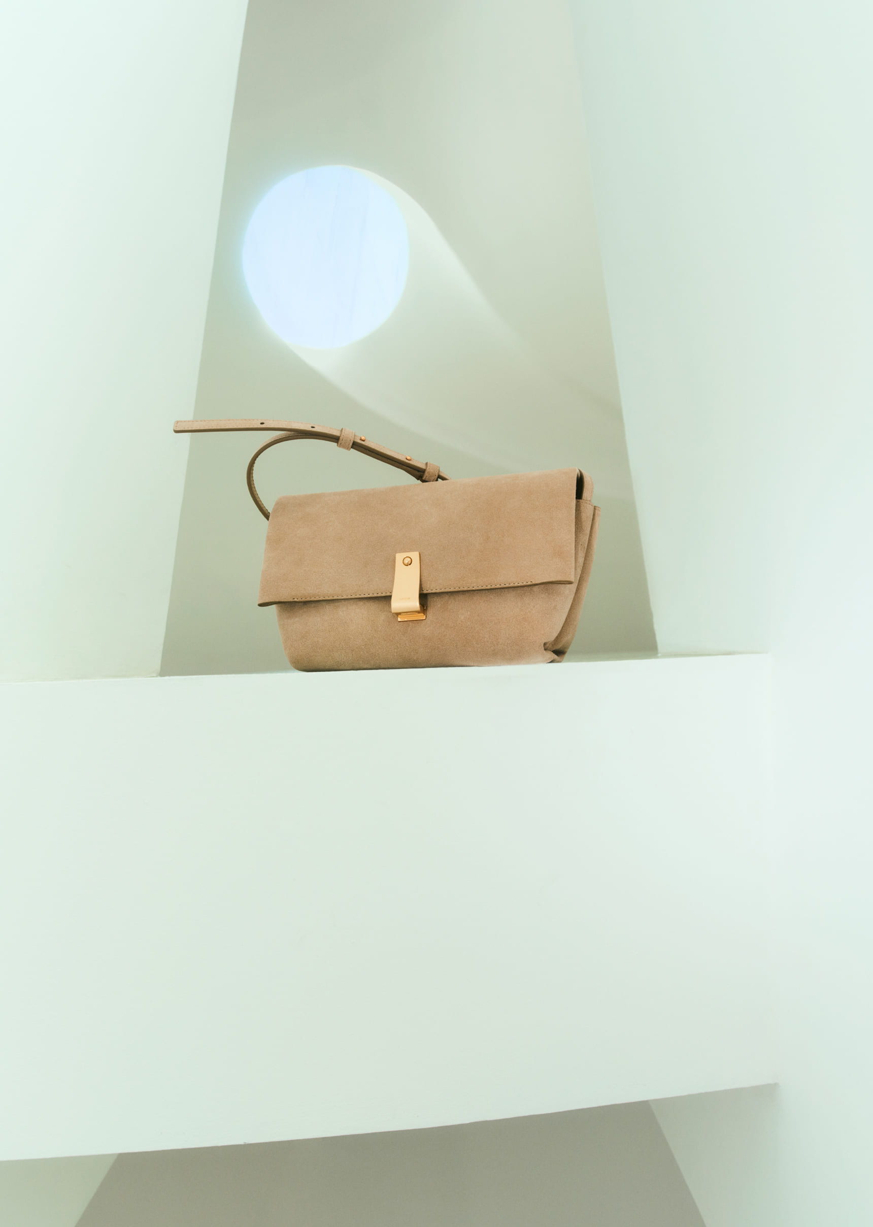 A beige handbag displayed on a minimalistic white shelf.