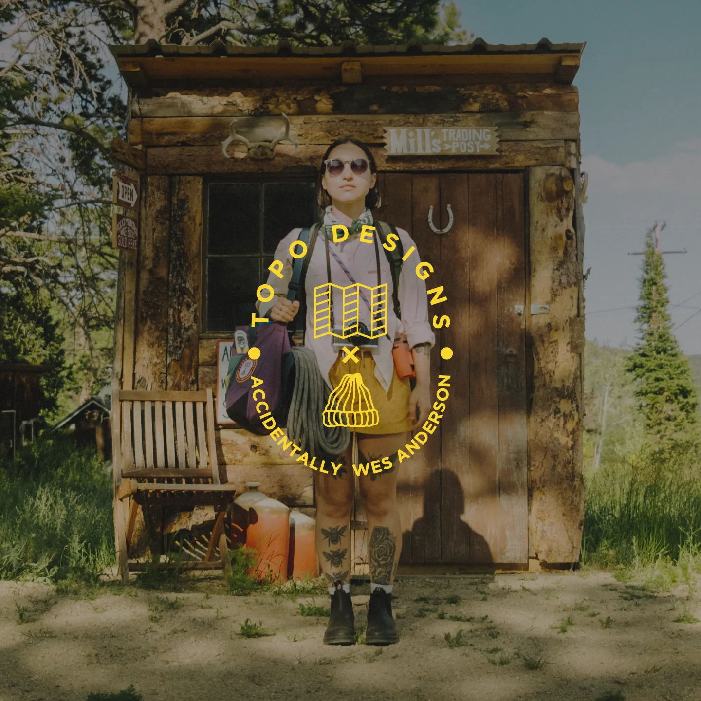 Person standing in front of a rustic cabin, wearing outdoor clothing and sunglasses.