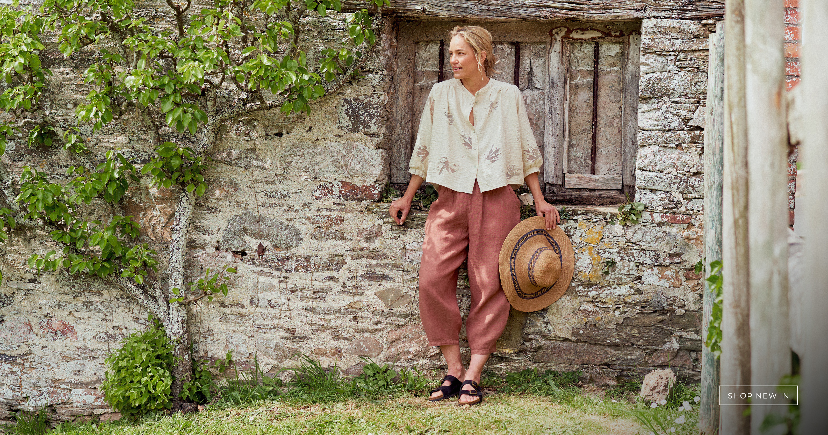 Woman wearing rust linen trousers and white textured shirt against a rustic wall 