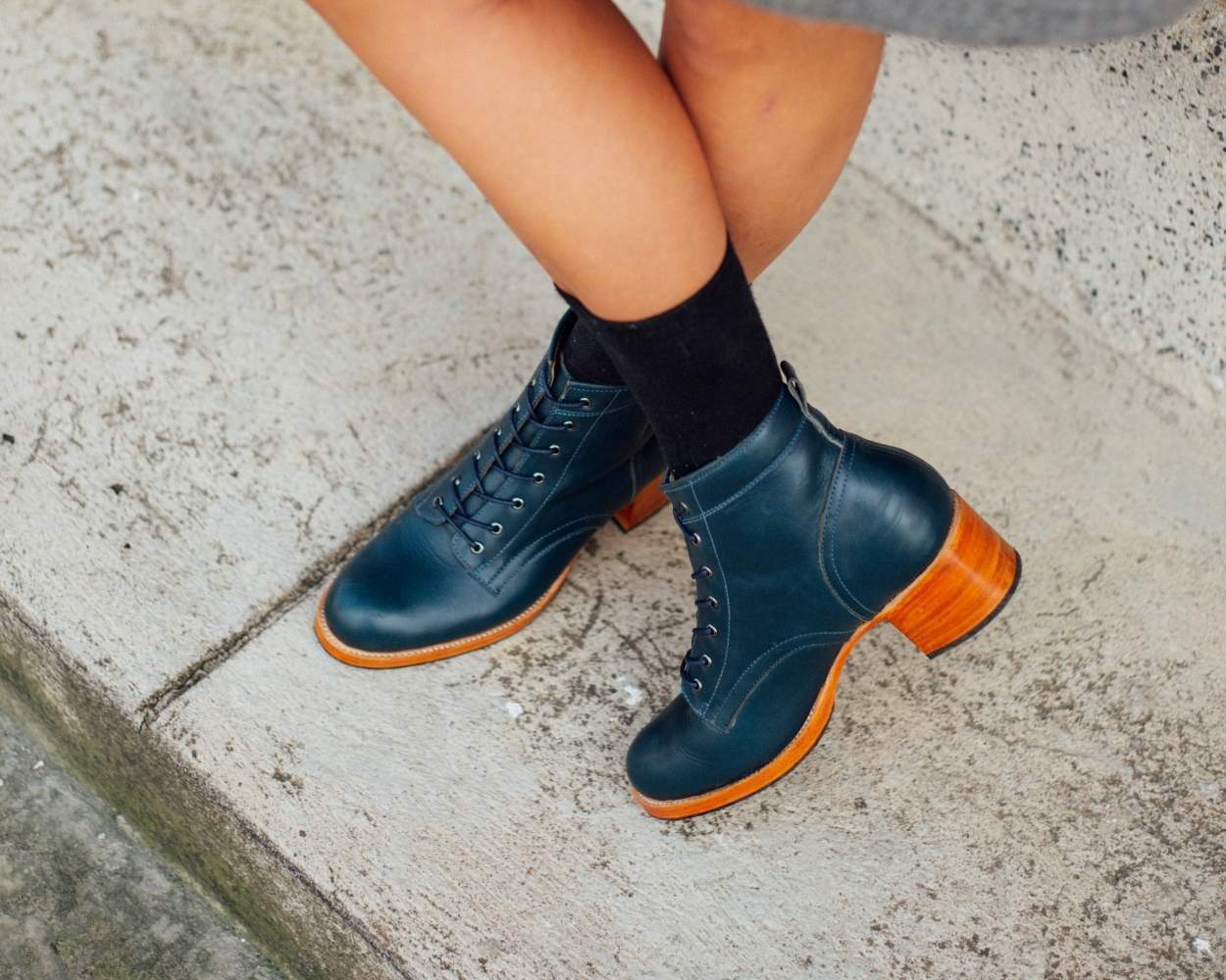 A close-up lifestyle shot from the calves down of a person standing with legs crossed on a concrete step. They are wearing mid-calf black socks and a pair of handmade denim blue leather lace-up boots featuring a rounded toe, matching blue laces, and a prominent, light-colored stacked block heel.