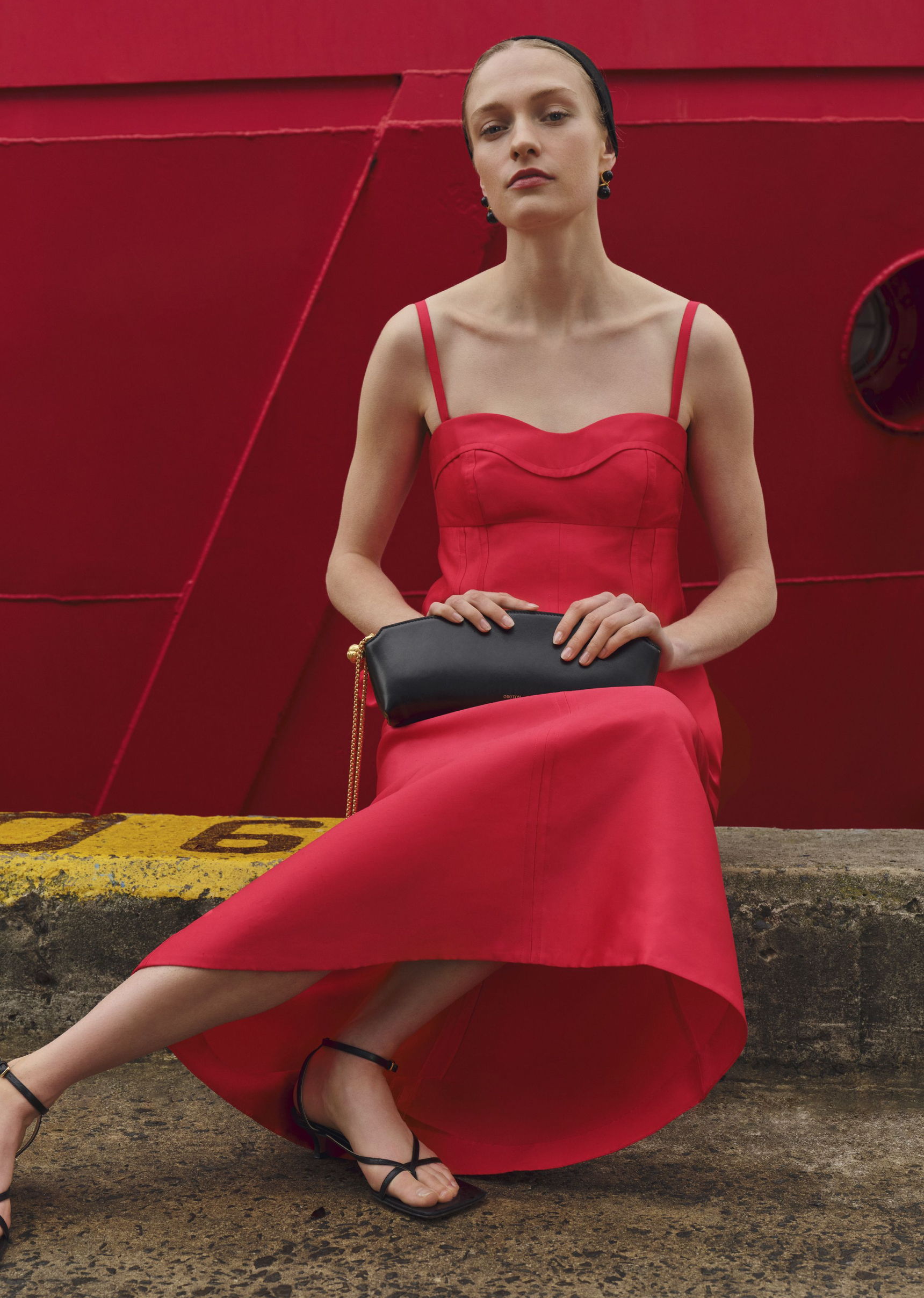 Model in a red dress sitting with a clutch against a red background.