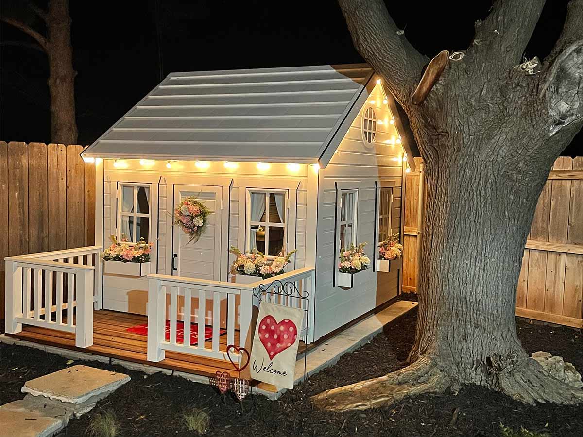 Wooden playhouse in white color in a corner of a backyard with lights next to a tree by WholeWoodPlayhouses