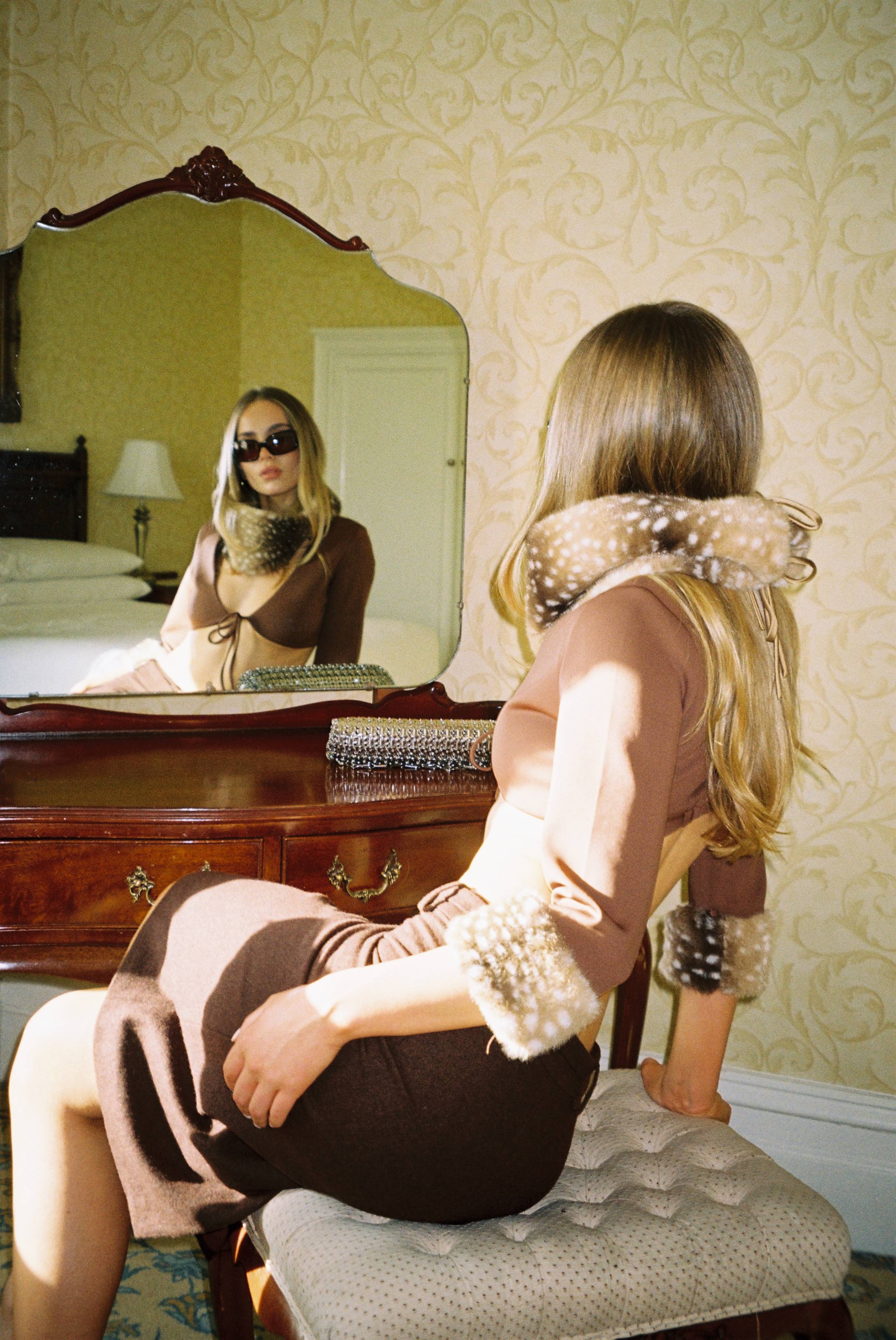 Woman in stylish outfit sitting at a vanity, looking into a mirror.