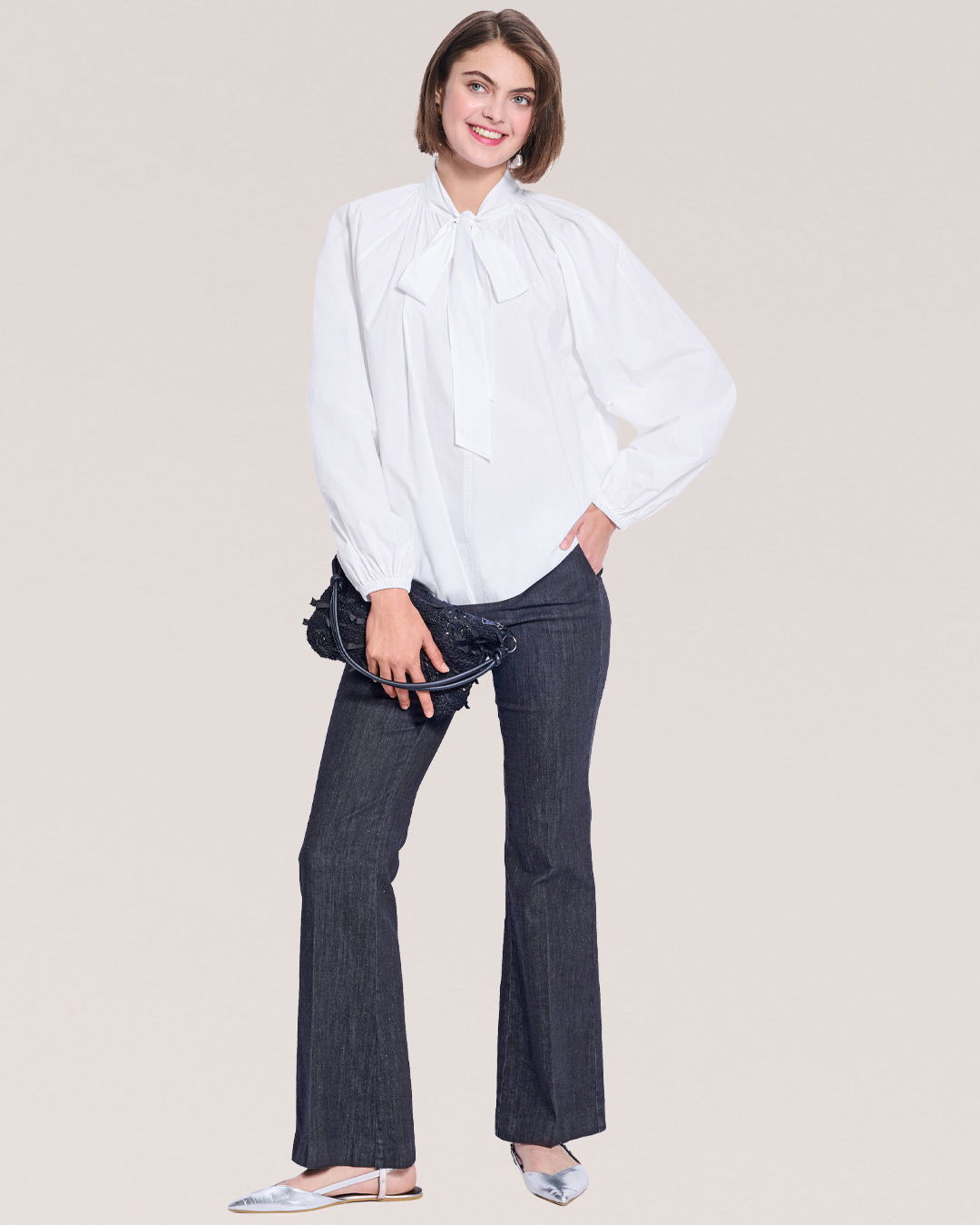 Woman in a white blouse and dark jeans, posing with a clutch bag.