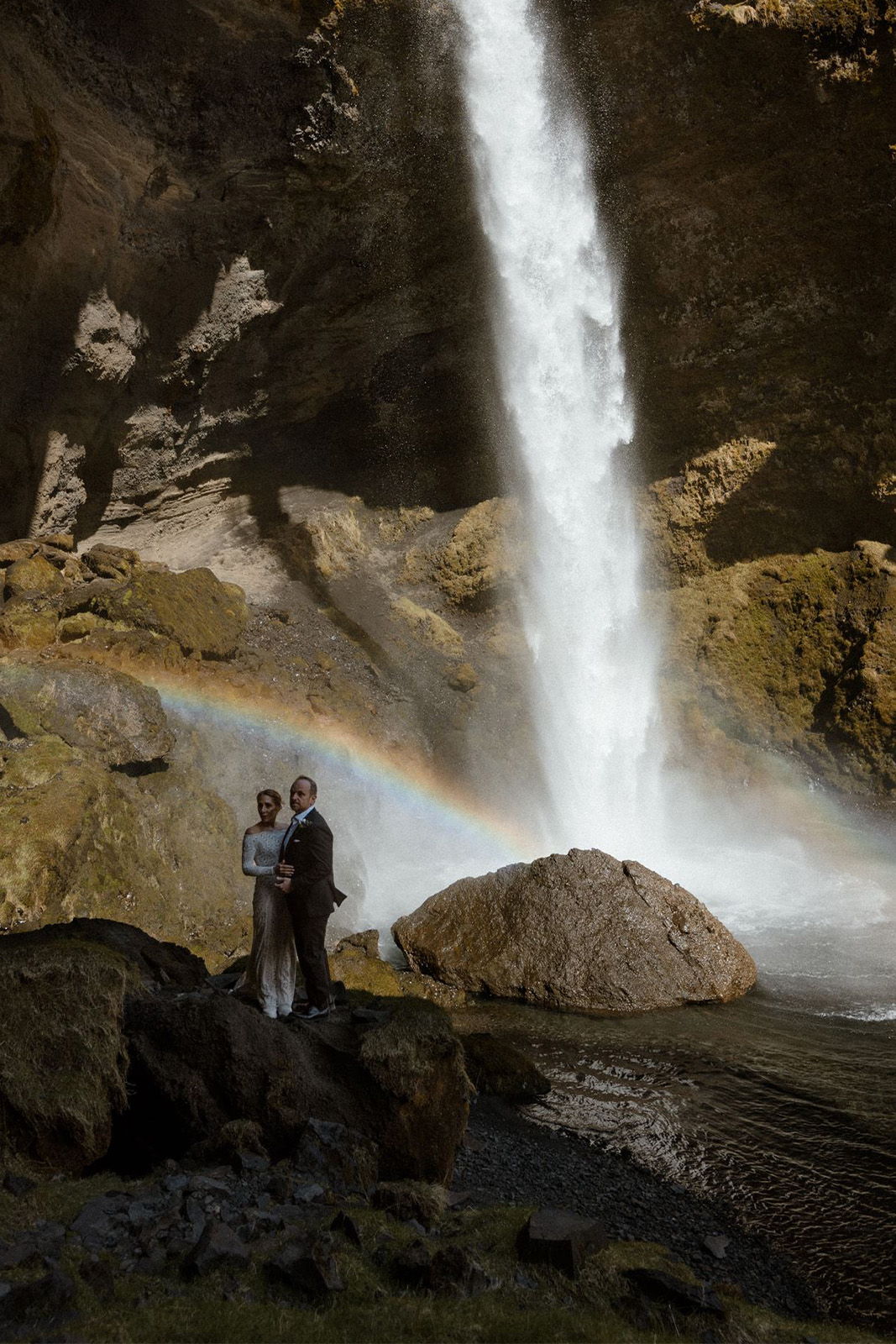 A couple standing by a waterfall with a rainbow in the background.