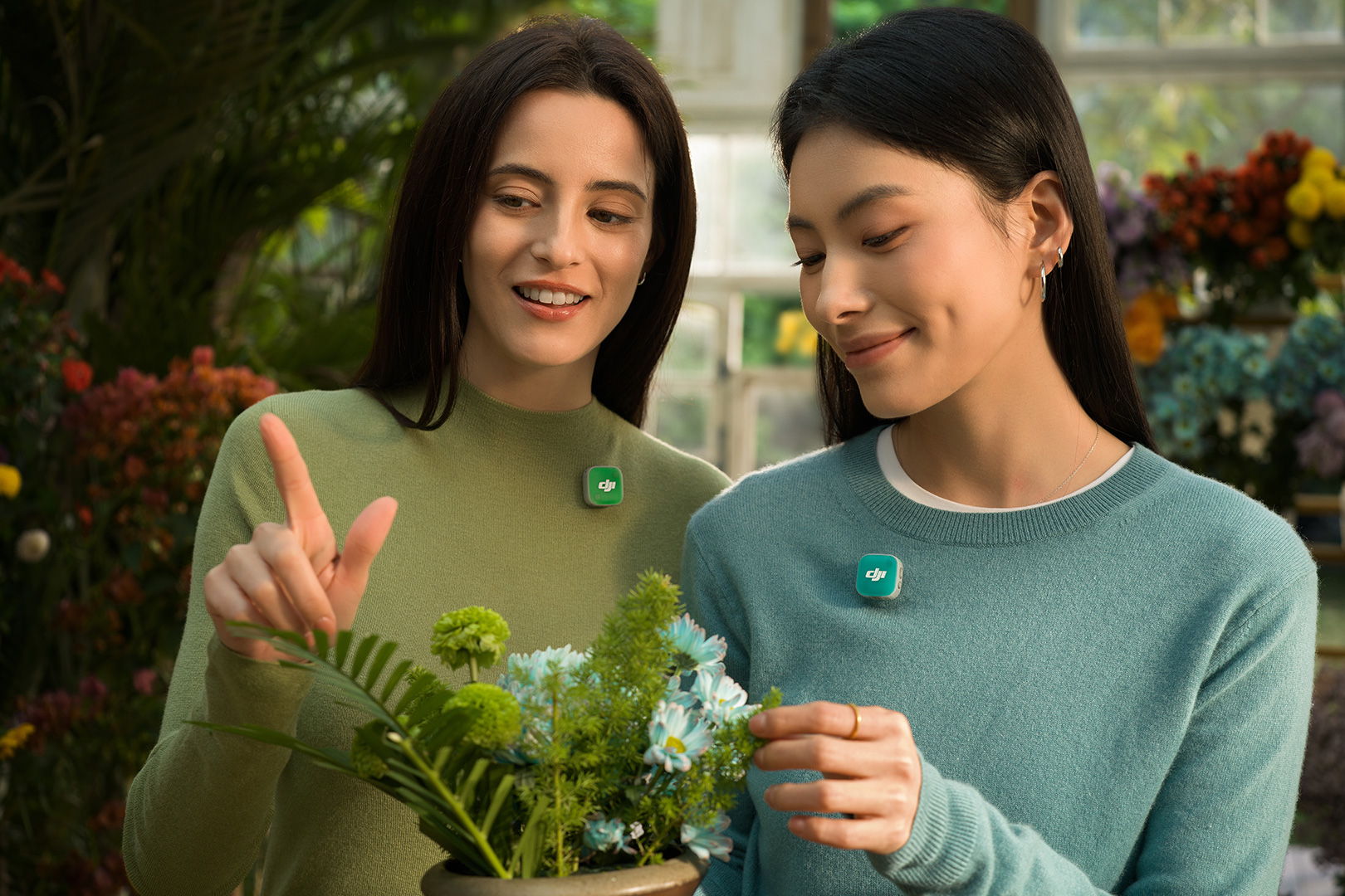 Two women smiling and interacting while tending to plants in a greenhouse, they're using DJI Mic Mini 2 to record their audio with faceplates matching their sweaters.
