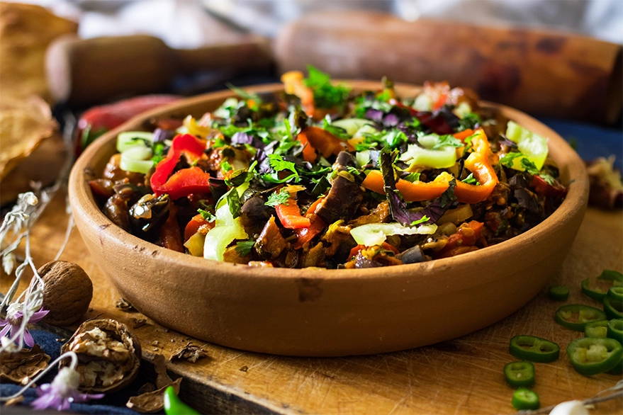 Georgian ajapsandali in a clay bowl with roasted eggplants, peppers, onions, garnished with basil on a wooden table, a vegetarian staple of Suneli Valley cuisine.