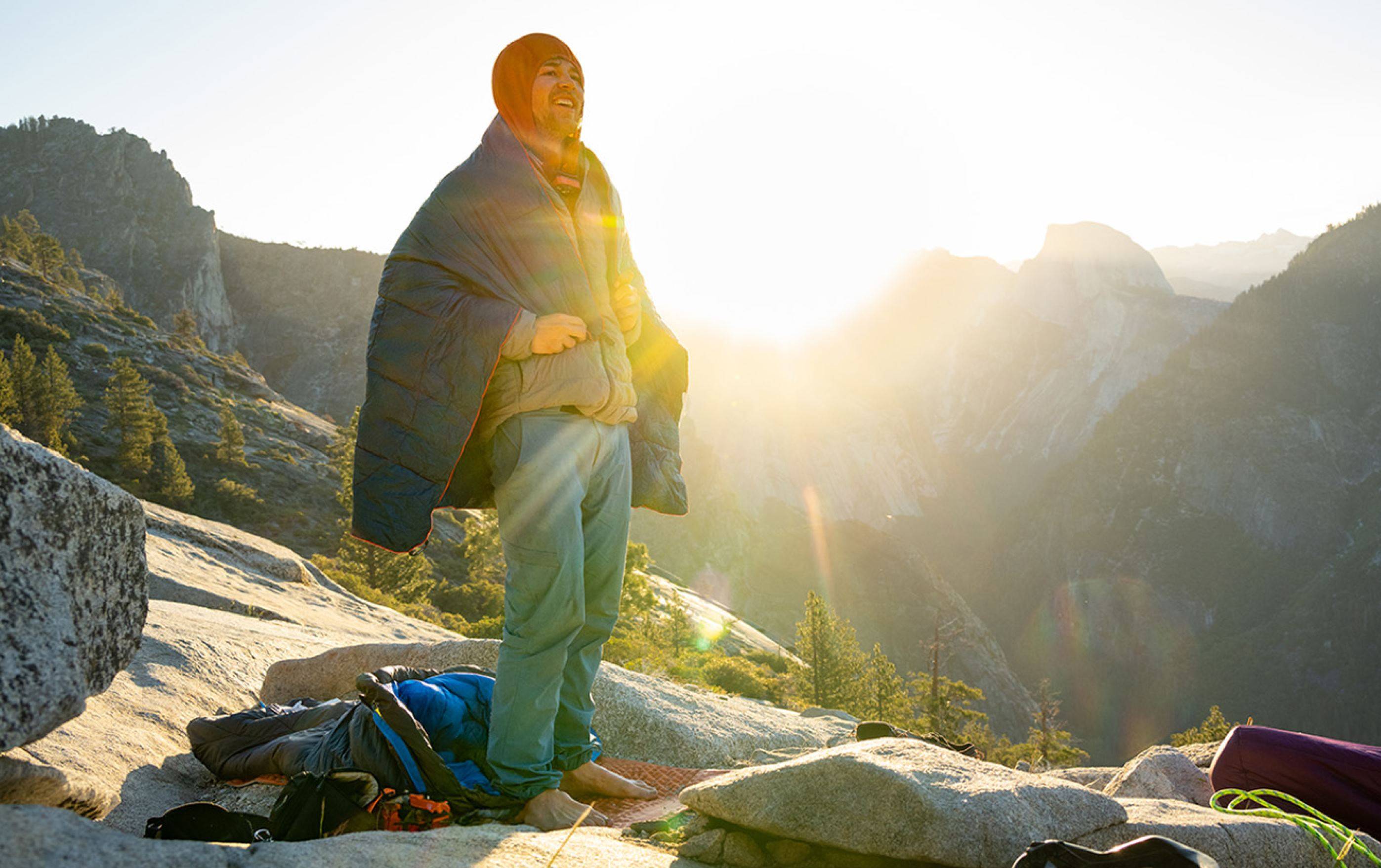 A person on a mountain at sunrise, wearing a Rumpl around his shoulders