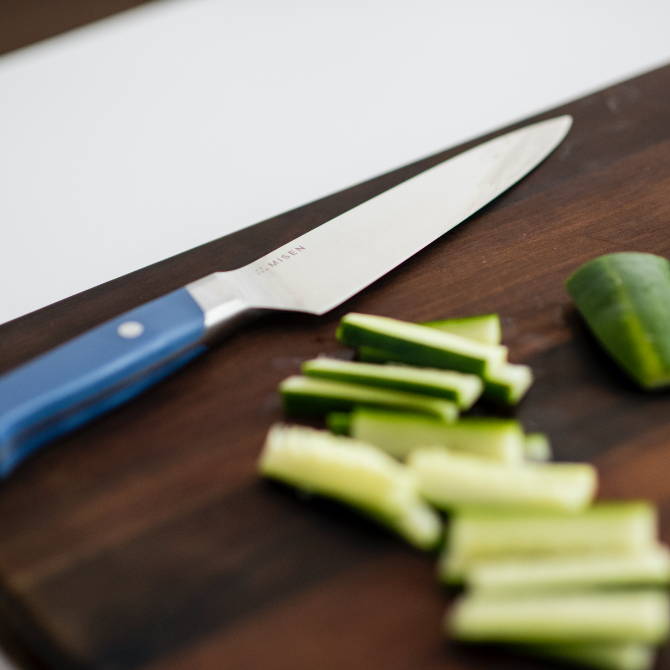 A Blue Misen Utility Knife resting on a dark wood cutting board, next to thinly-sliced cucumber sticks.