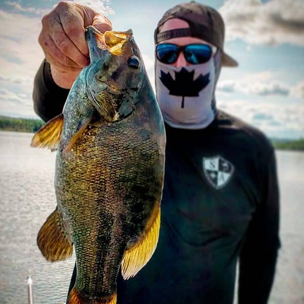 Mitch Dupuis holding a fish up to the camera while wearing an SA Company black performance shirt, sunglasses, hat and a black and white Canadian face shield over his face.