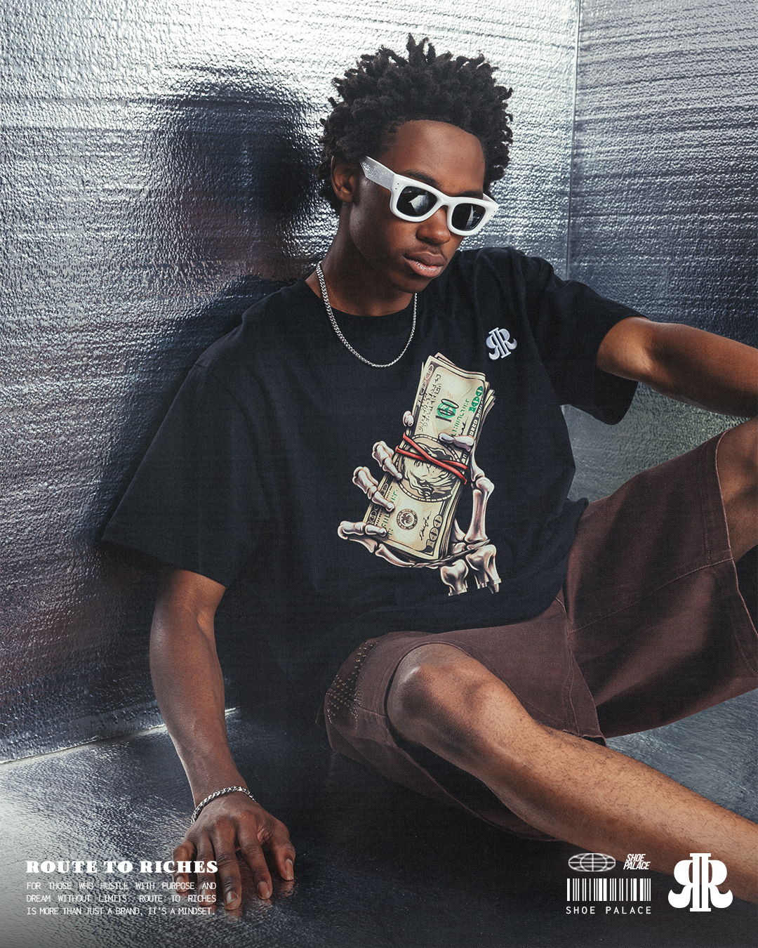 Young person wearing sunglasses and a graphic t-shirt, sitting against a wall.