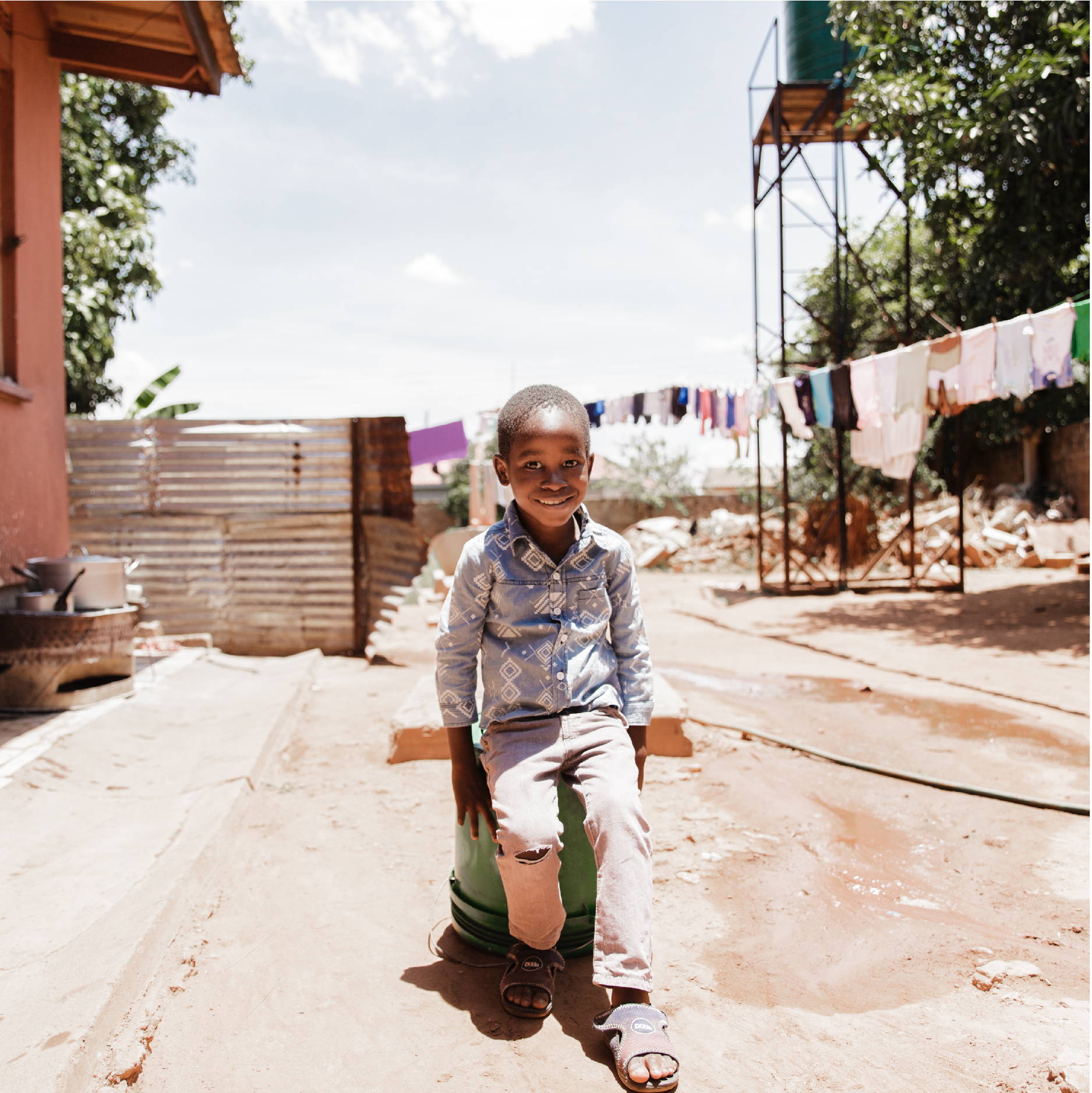 A young Zambian boy sits on a bucket outside his orphanage. The ground is very dry and clothes are hanging in the background