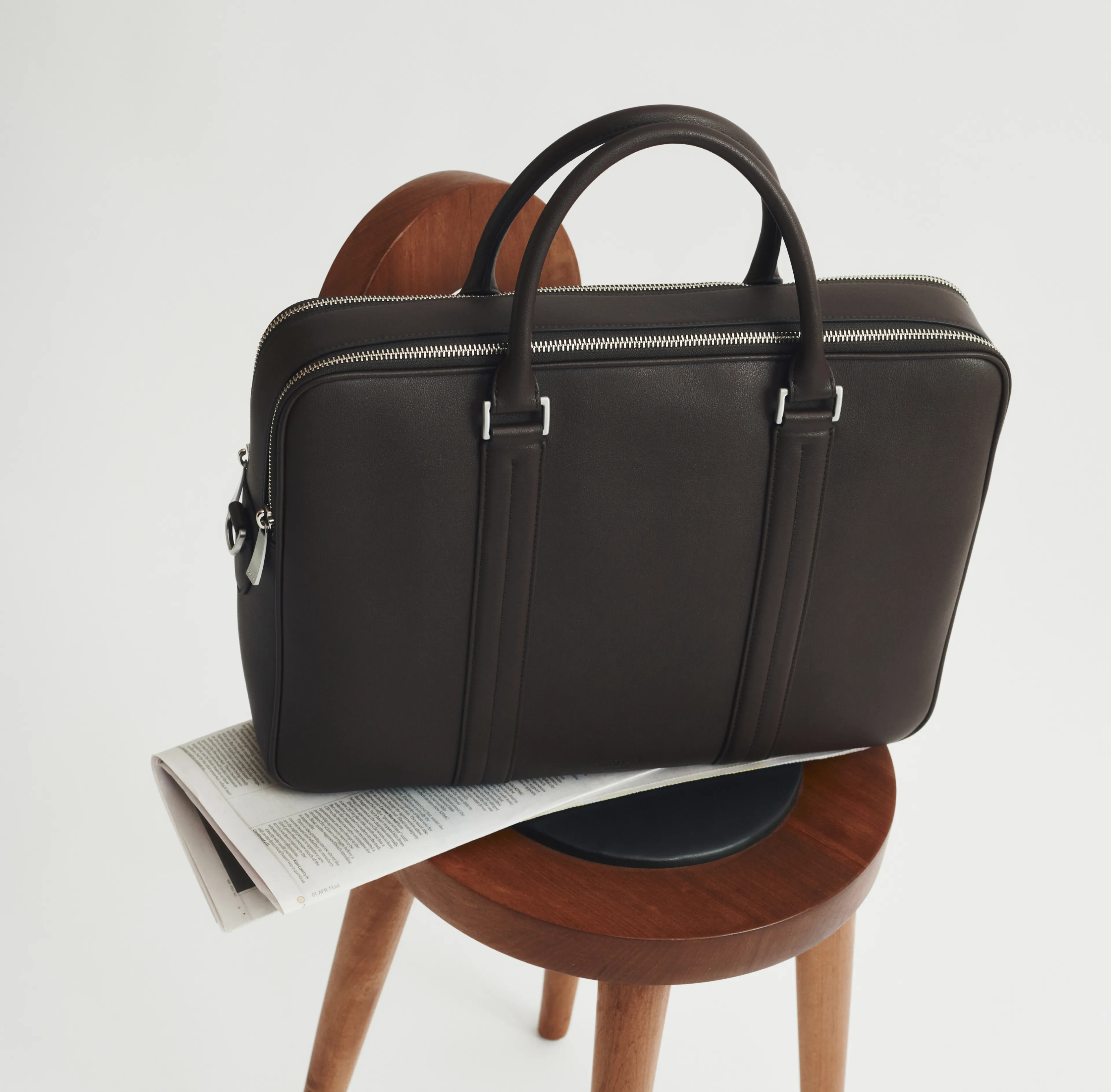 A brown leather bag resting on a wooden stool with a newspaper underneath.