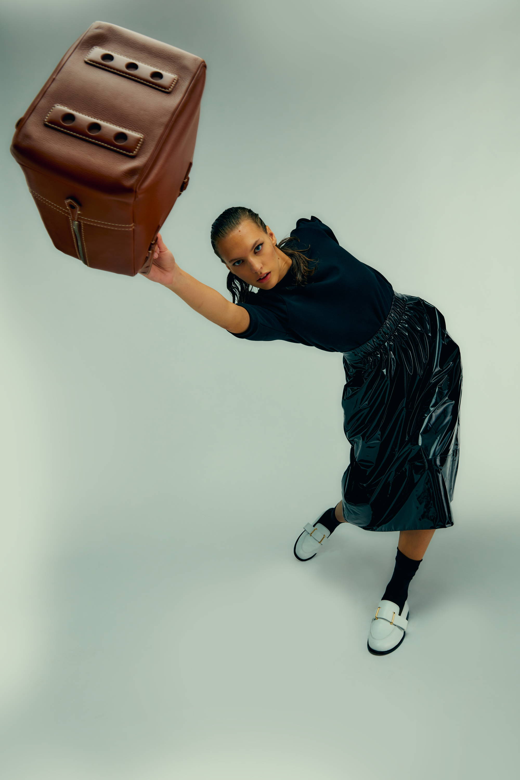 Person in a skirt lifting a large brown bag against a light background.