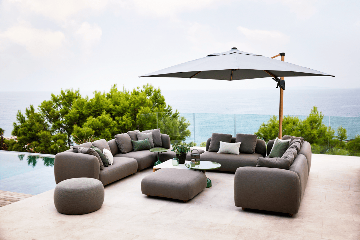 Large cantilever patio umbrella shading a modular gray outdoor sectional on a stone terrace beside a pool with an ocean view.