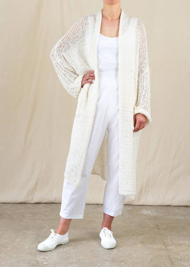 Model wearing long white crochet cardigan, white top, white trousers and white shoes.